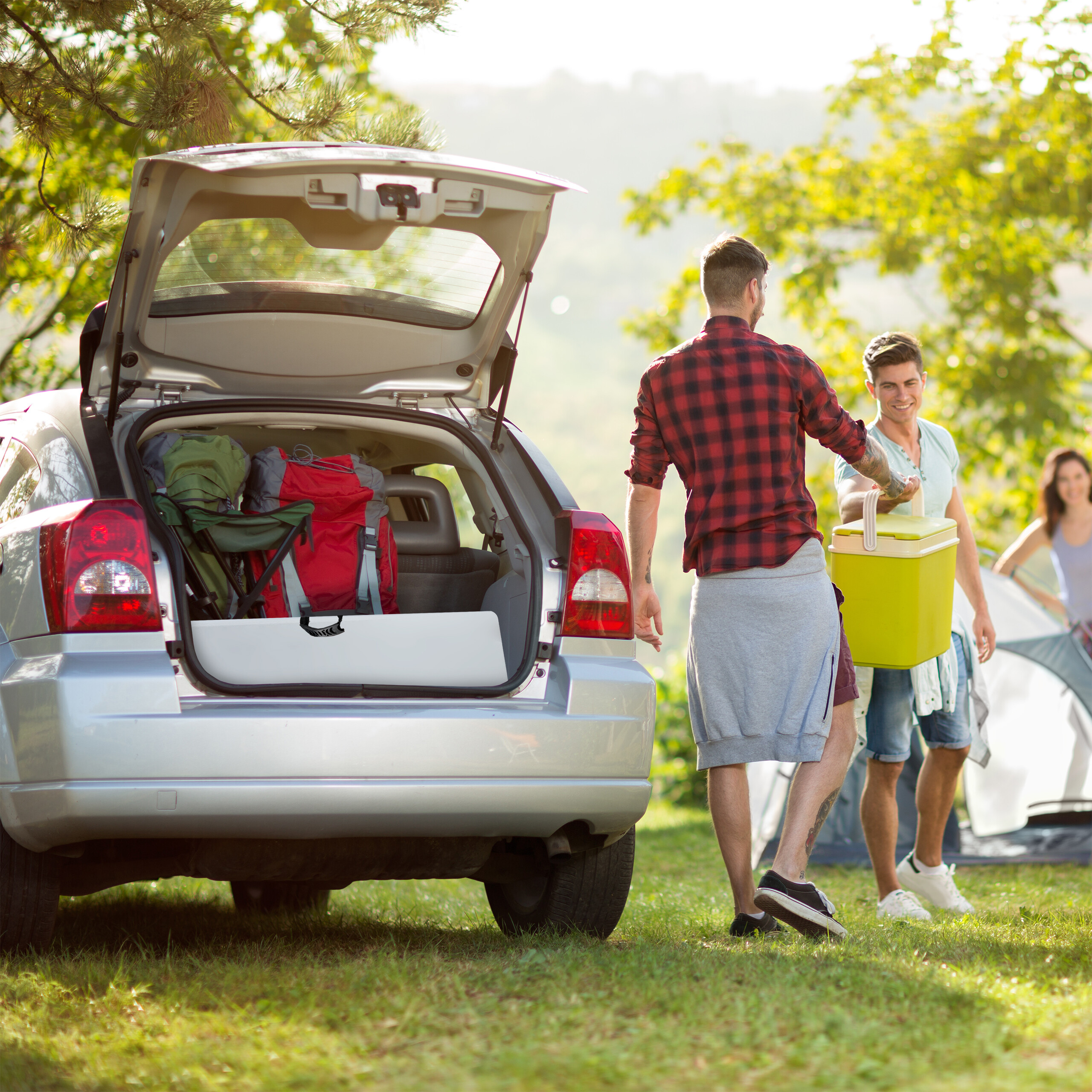 Zwei Männer packen eine Kühlbox aus dem Kofferraum eines silbernen Autos beim Camping.