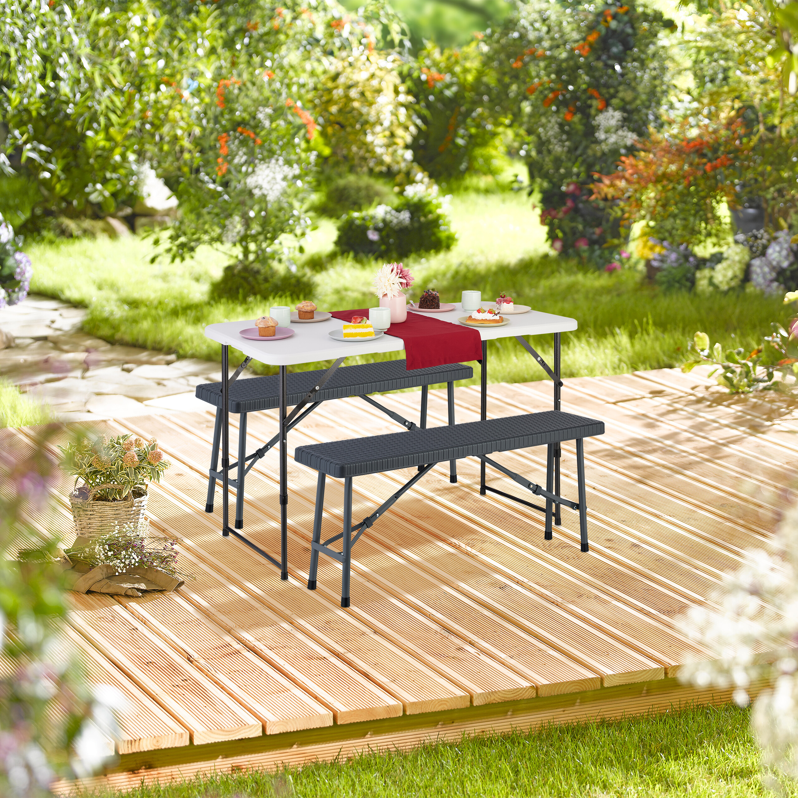 Ein weißer Tisch mit Bänken steht auf einer Holzterrasse im Garten, gedeckt mit Kuchen und Blumen.