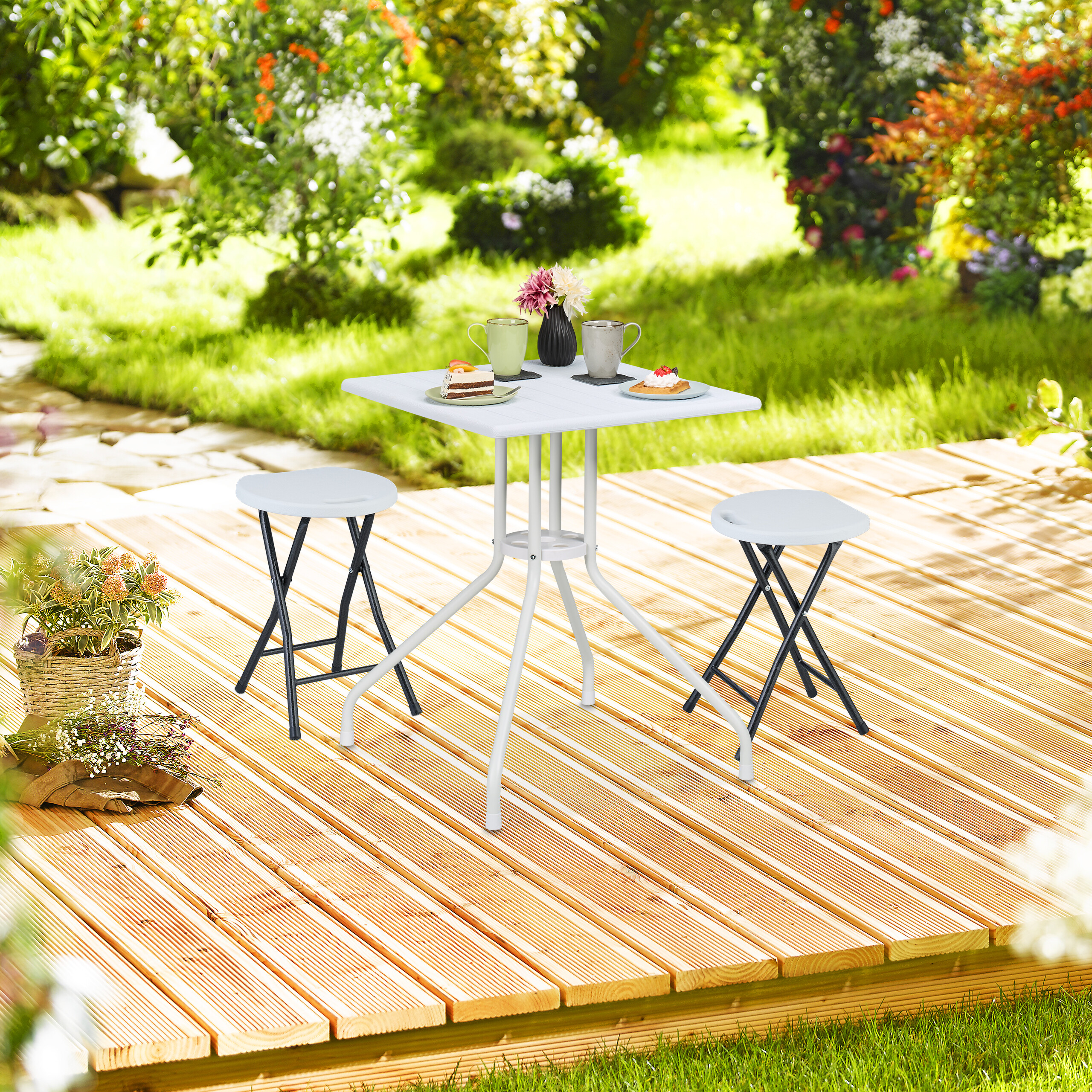Ein weißer Tisch mit zwei Hockern auf einer Holzterrasse im Garten, gedeckt mit Kuchen und Tassen.