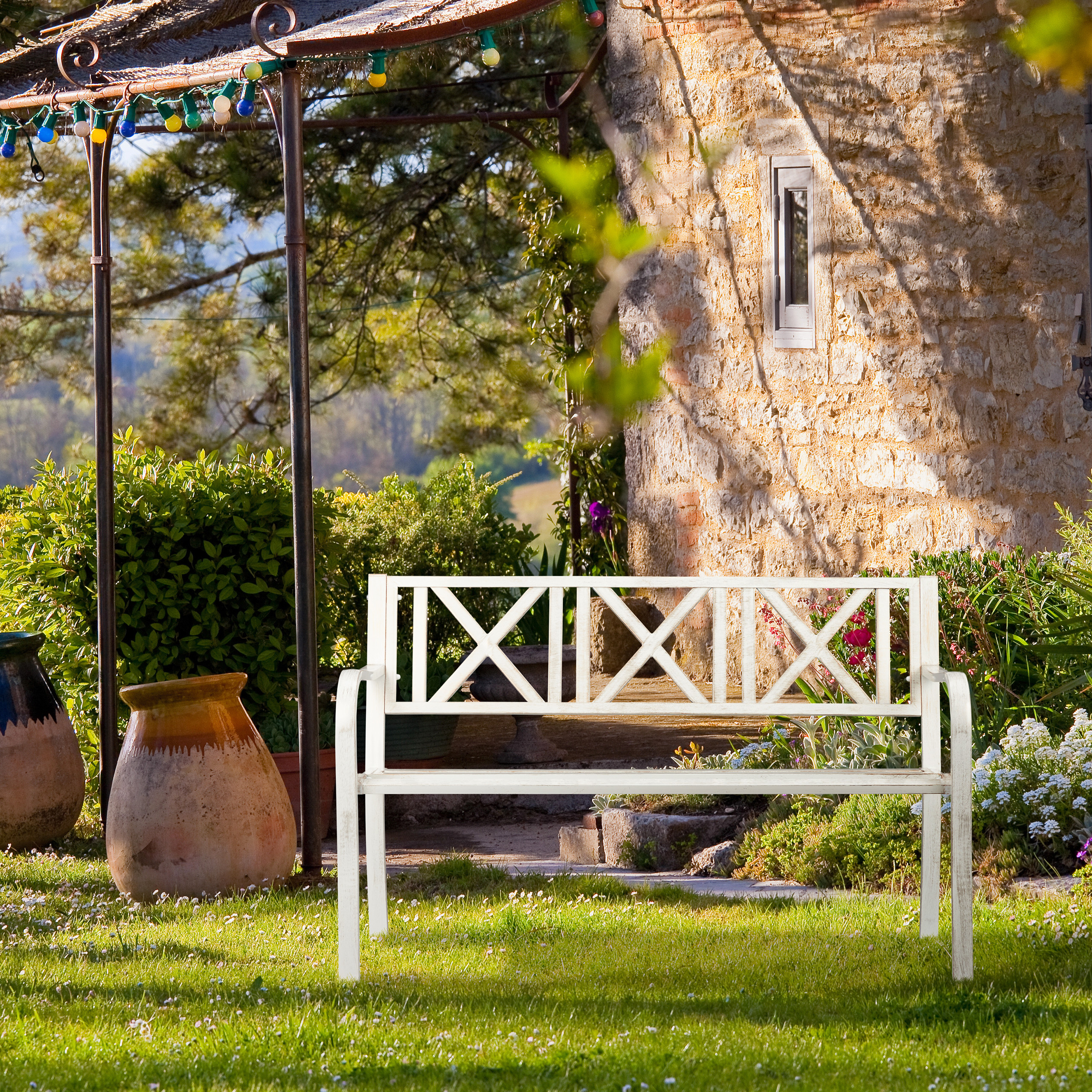 Eine weiße Bank im Garten vor einem Steinhaus mit Pergola und Pflanzen.