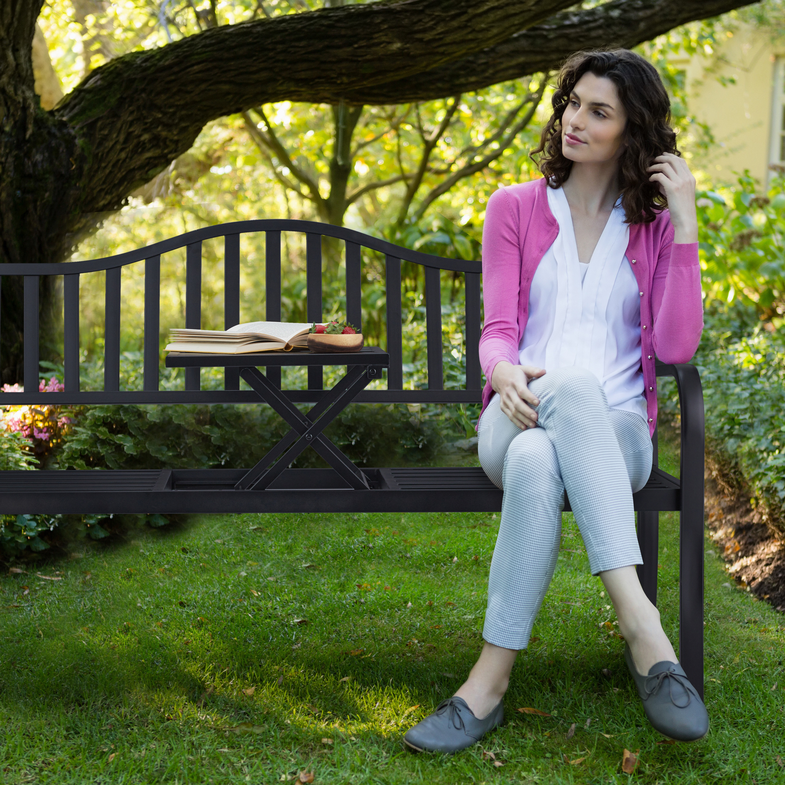 Eine Frau in rosa Jacke sitzt auf einer schwarzen Gartenbank im Park mit einem Buch und Früchten auf dem Klapptisch.