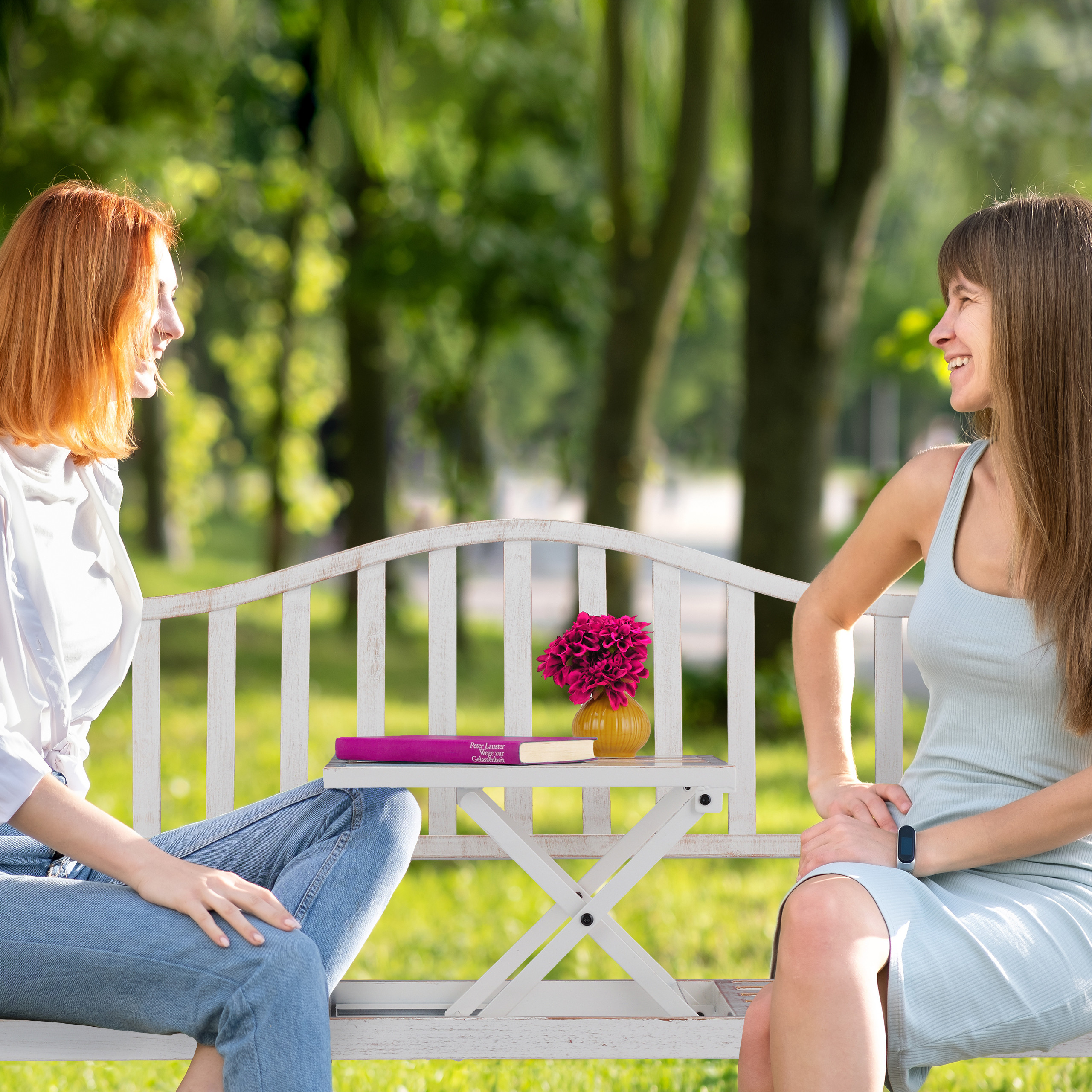 Zwei Frauen sitzen auf einer weißen Parkbank und unterhalten sich in einer grünen Parklandschaft.