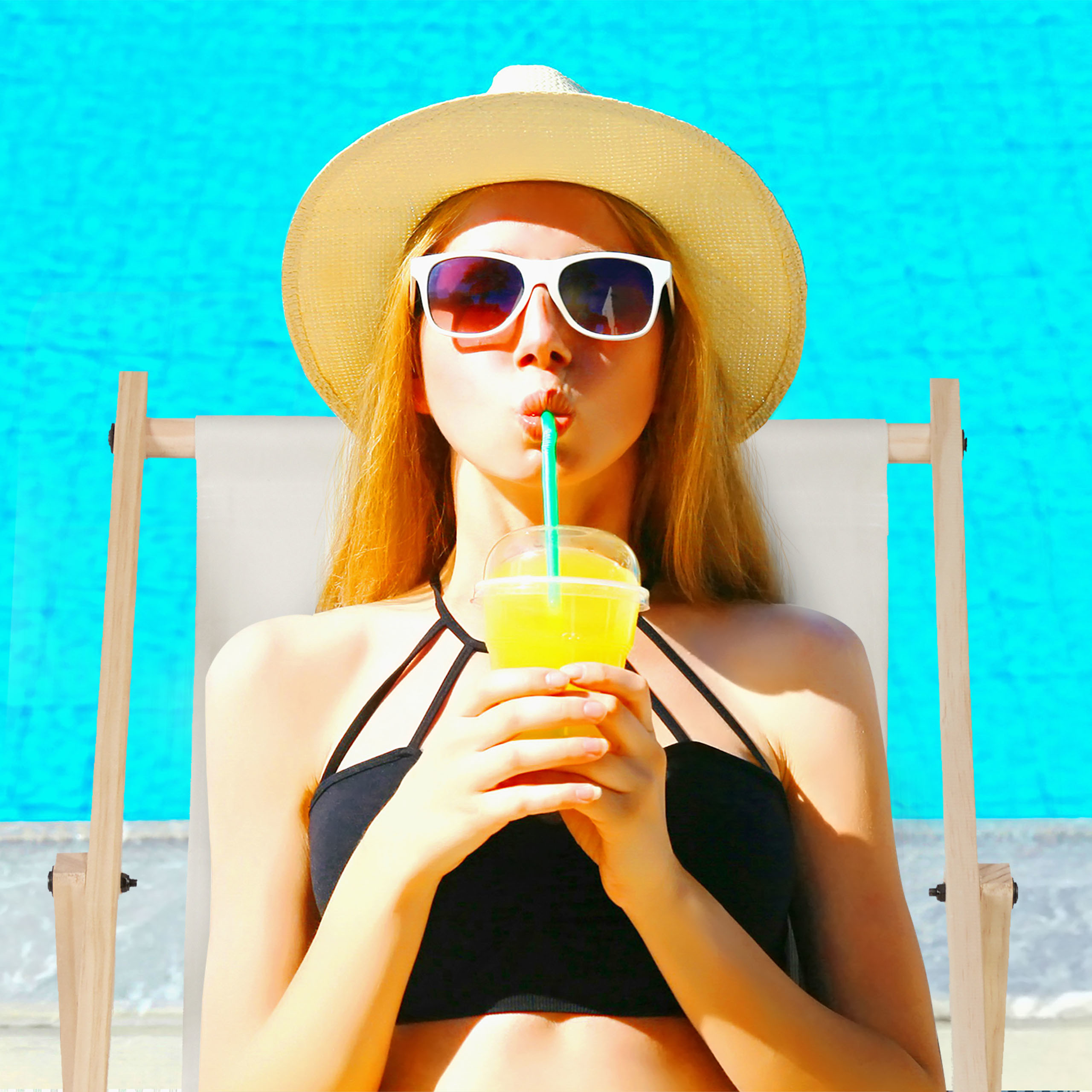Eine Frau mit Sonnenhut und Brille entspannt auf einer Liege am Pool und trinkt Saft mit Strohhalm.