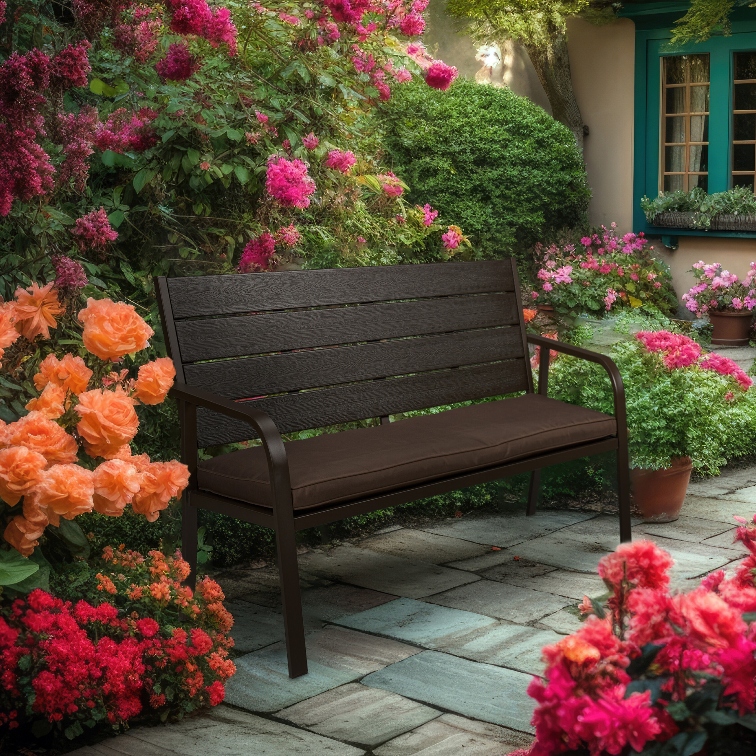 Eine braune Gartenbank mit Kissen steht auf einer Terrasse, umgeben von bunten Blumen und grünen Pflanzen.