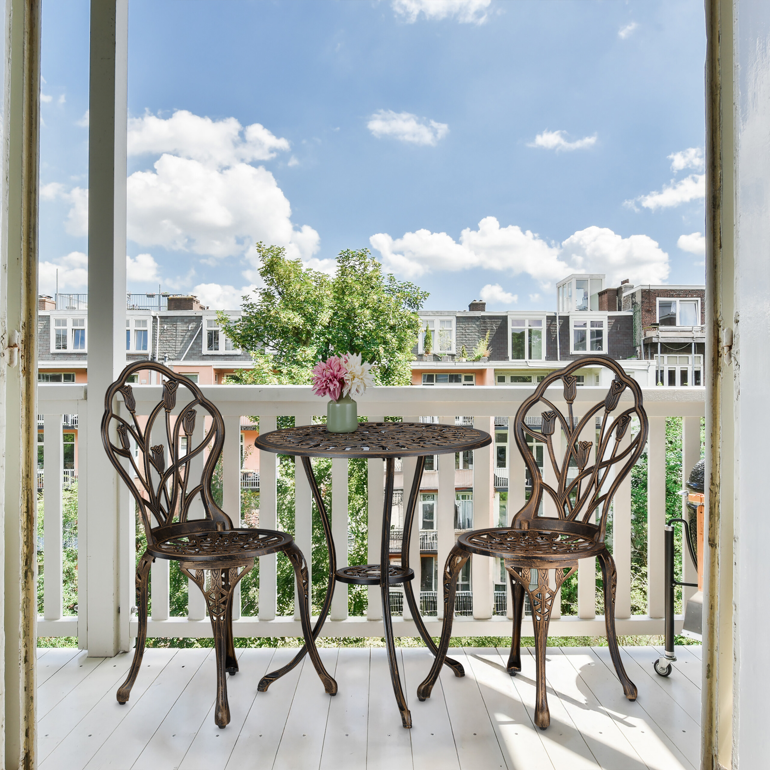 Ein kleiner Balkon mit Metalltisch, zwei Stühlen, Blumen und Blick auf Gebäude und blauen Himmel mit Wolken.