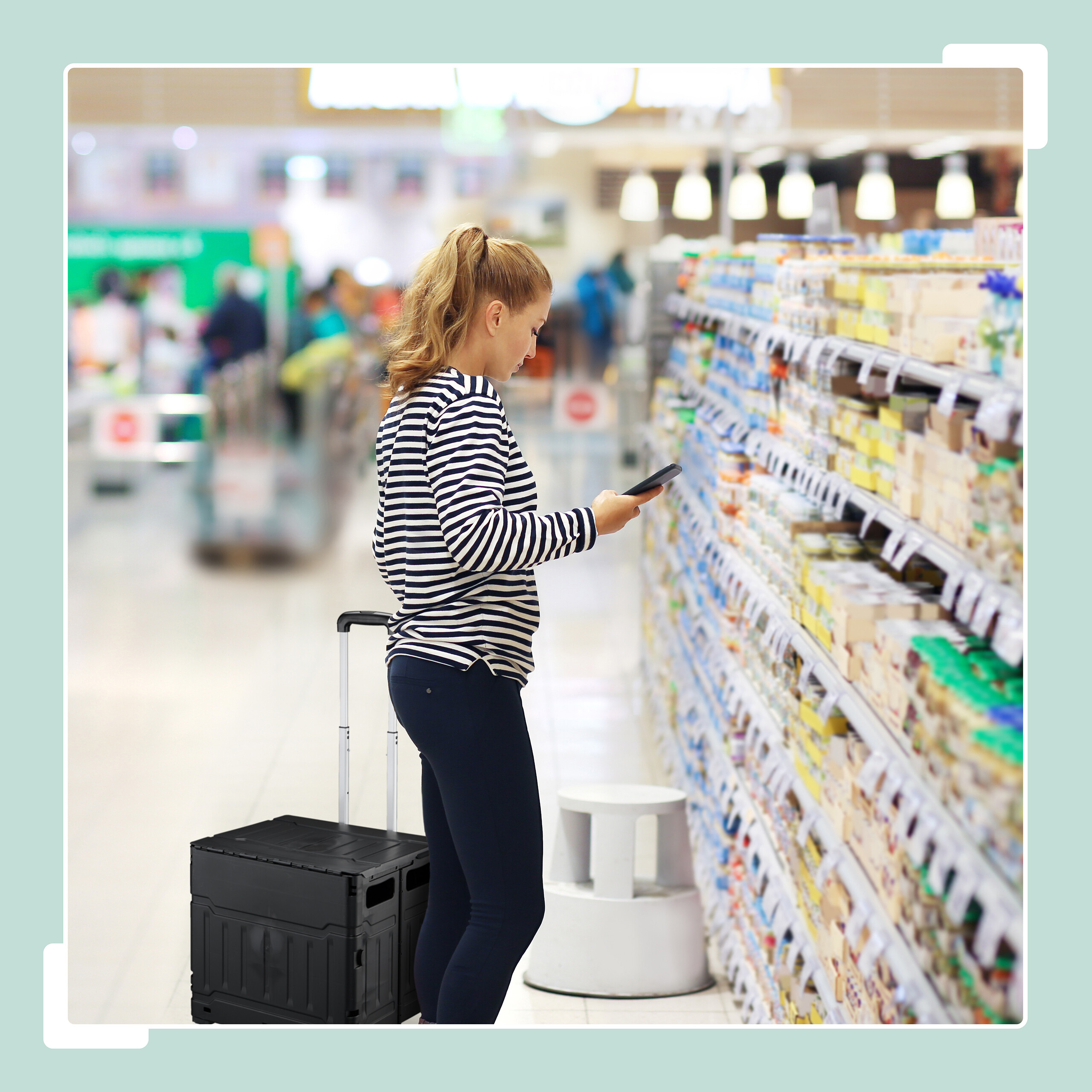 Eine blonde Frau im gestreiften Shirt schaut mit Koffer aufs Smartphone im Gang eines belebten Supermarkts.