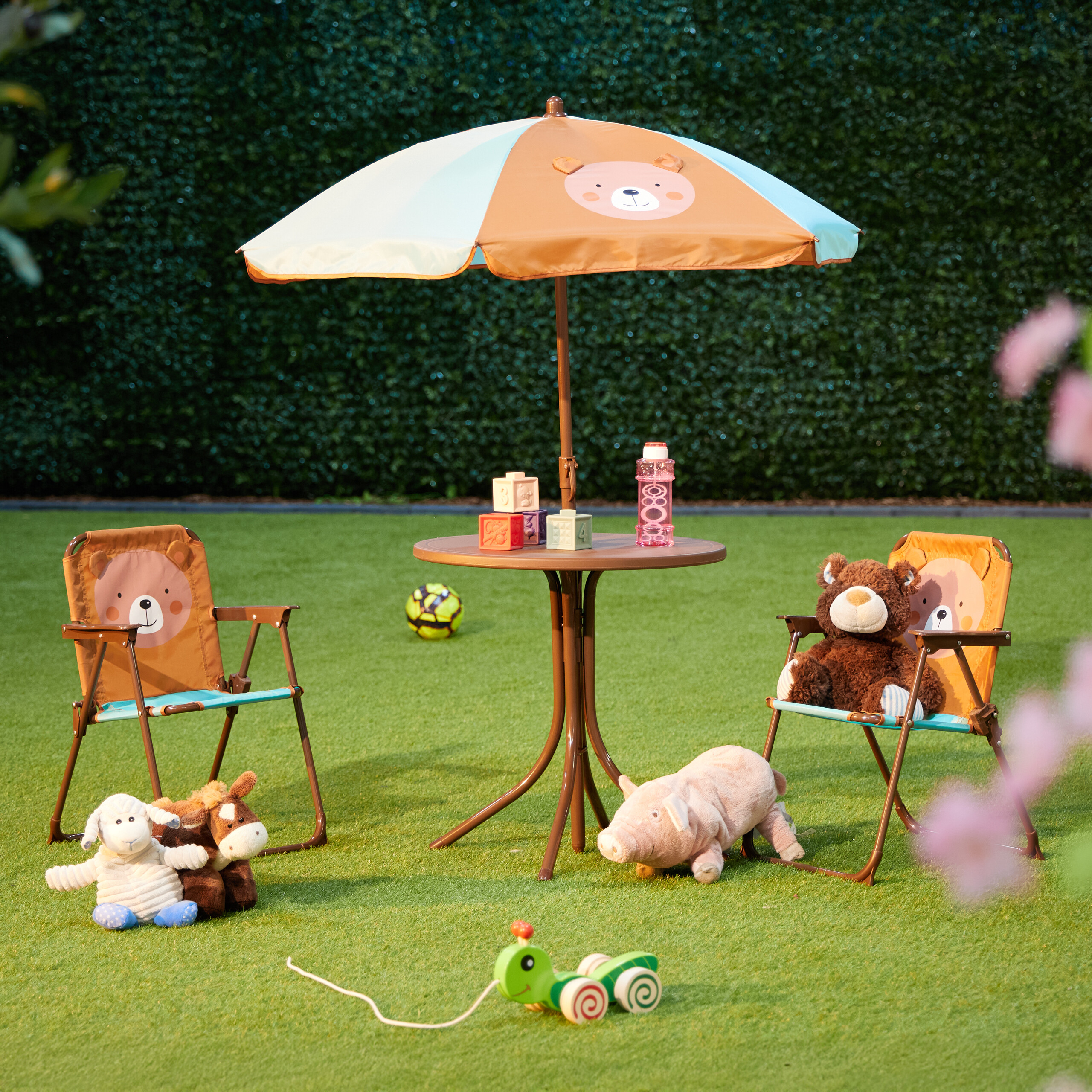 Ein Kindertisch mit Regenschirm, zwei Stühlen und Plüschtieren auf einer grünen Wiese vor einer Hecke.