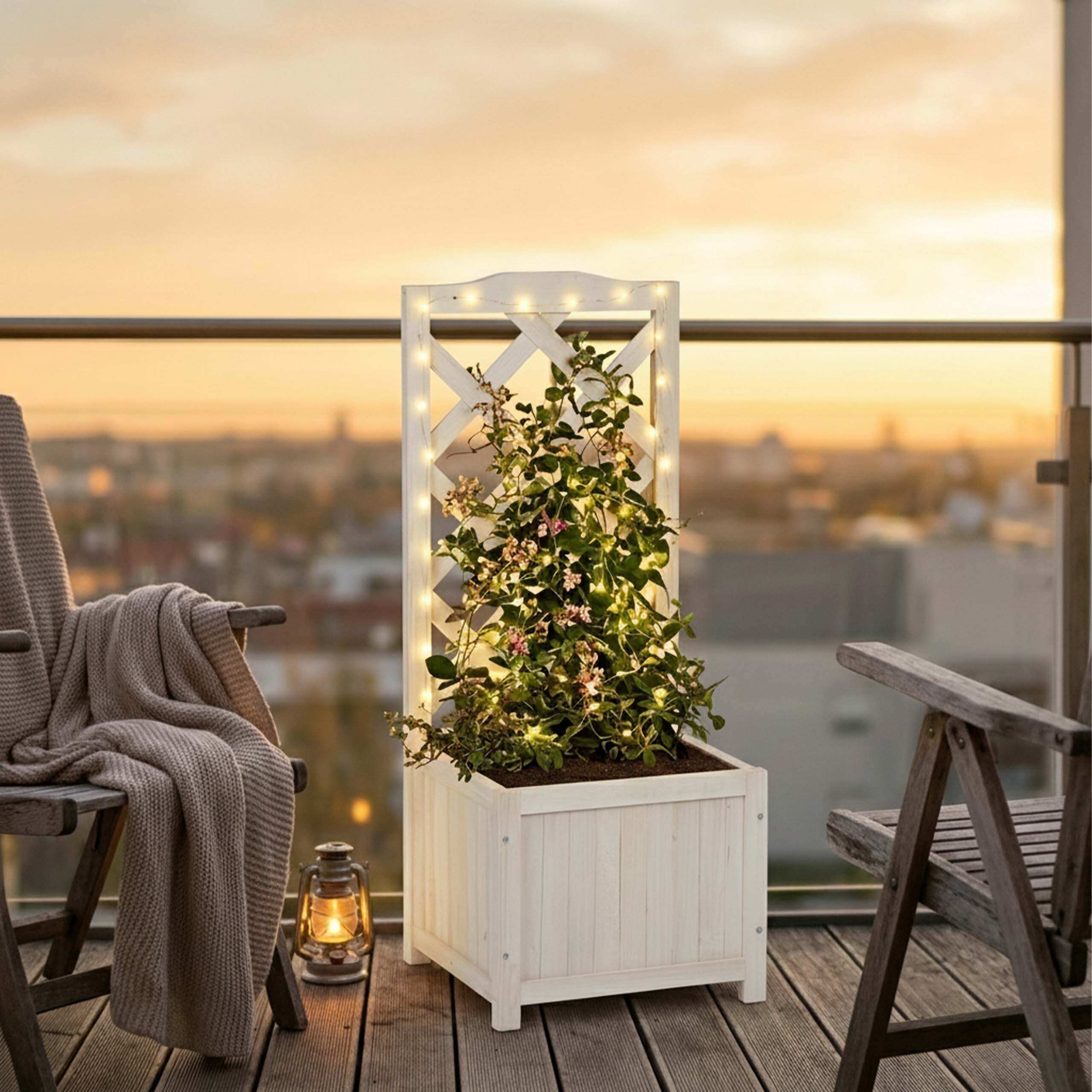 Ein weißer Holzblumenkübel mit Spalier, Lichterkette und Pflanzen steht bei Sonnenuntergang auf einem Balkon.