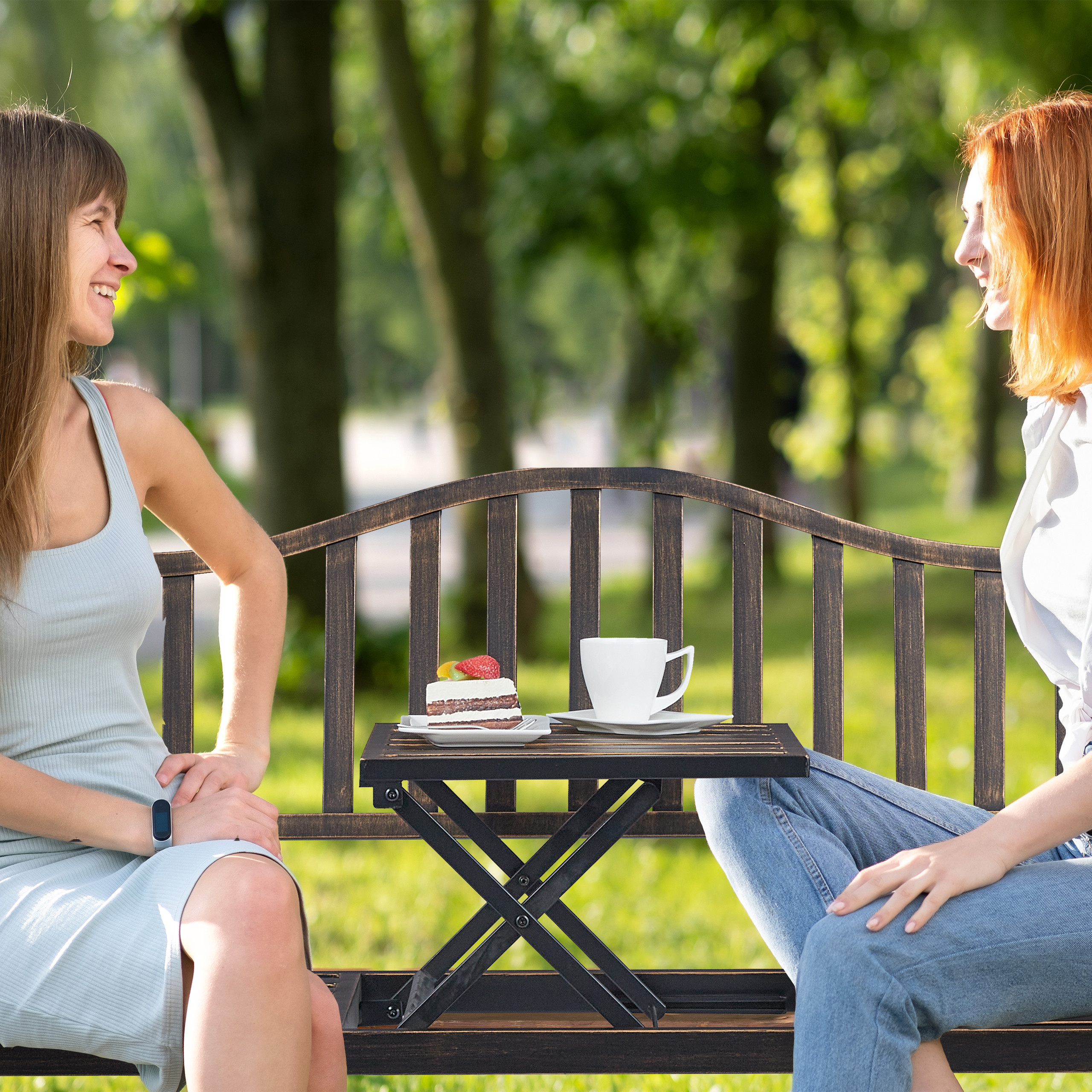Zwei Frauen sitzen auf einer Parkbank, zwischen ihnen steht ein kleiner Tisch mit Kuchen und einer Kaffeetasse.