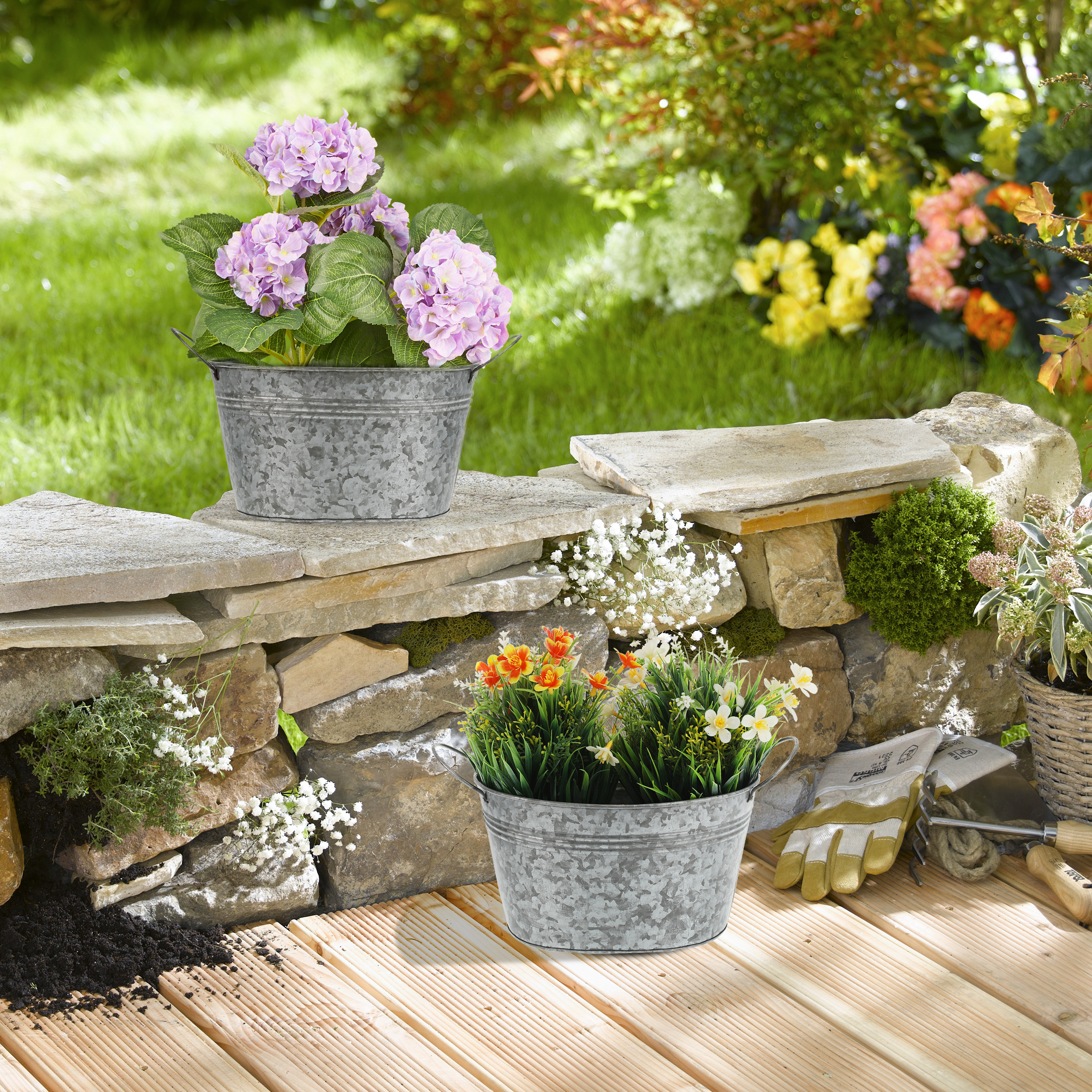 Zwei Zinkeimer mit rosa Hortensien und bunten Frühlingsblumen stehen auf einer Steinmauer im Garten.