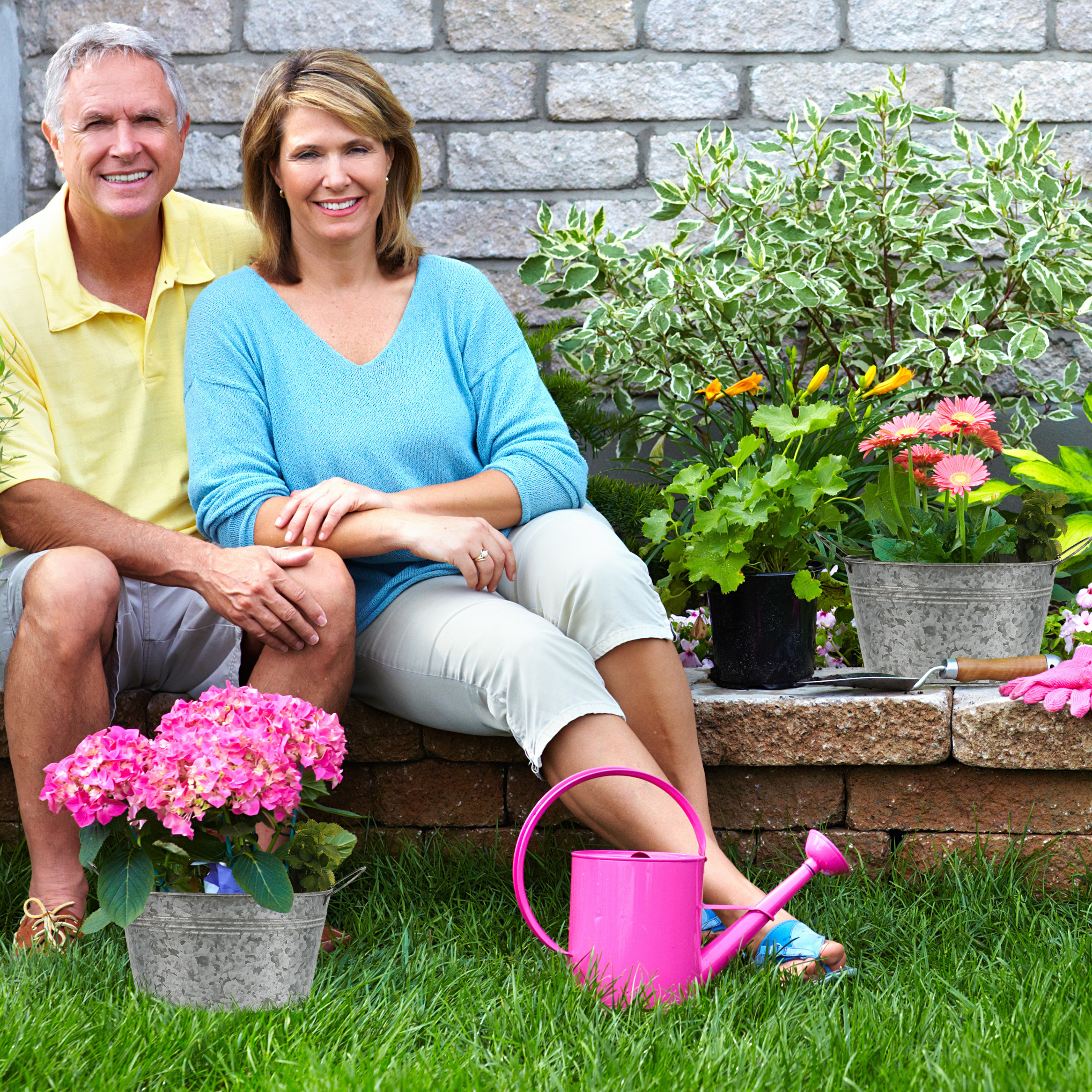 Glückliches älteres Paar sitzt vor einer Steinmauer im Garten mit Blumen und einer rosafarbenen Gießkanne.