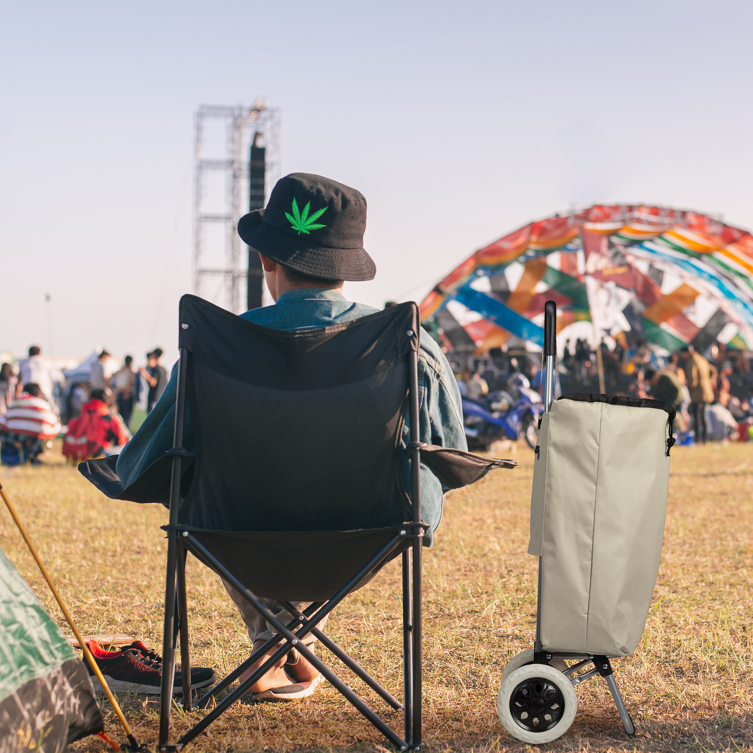 Eine Person sitzt auf einem Campingstuhl bei einem Festival mit einer Karre daneben und einer Bühne im Hintergrund.