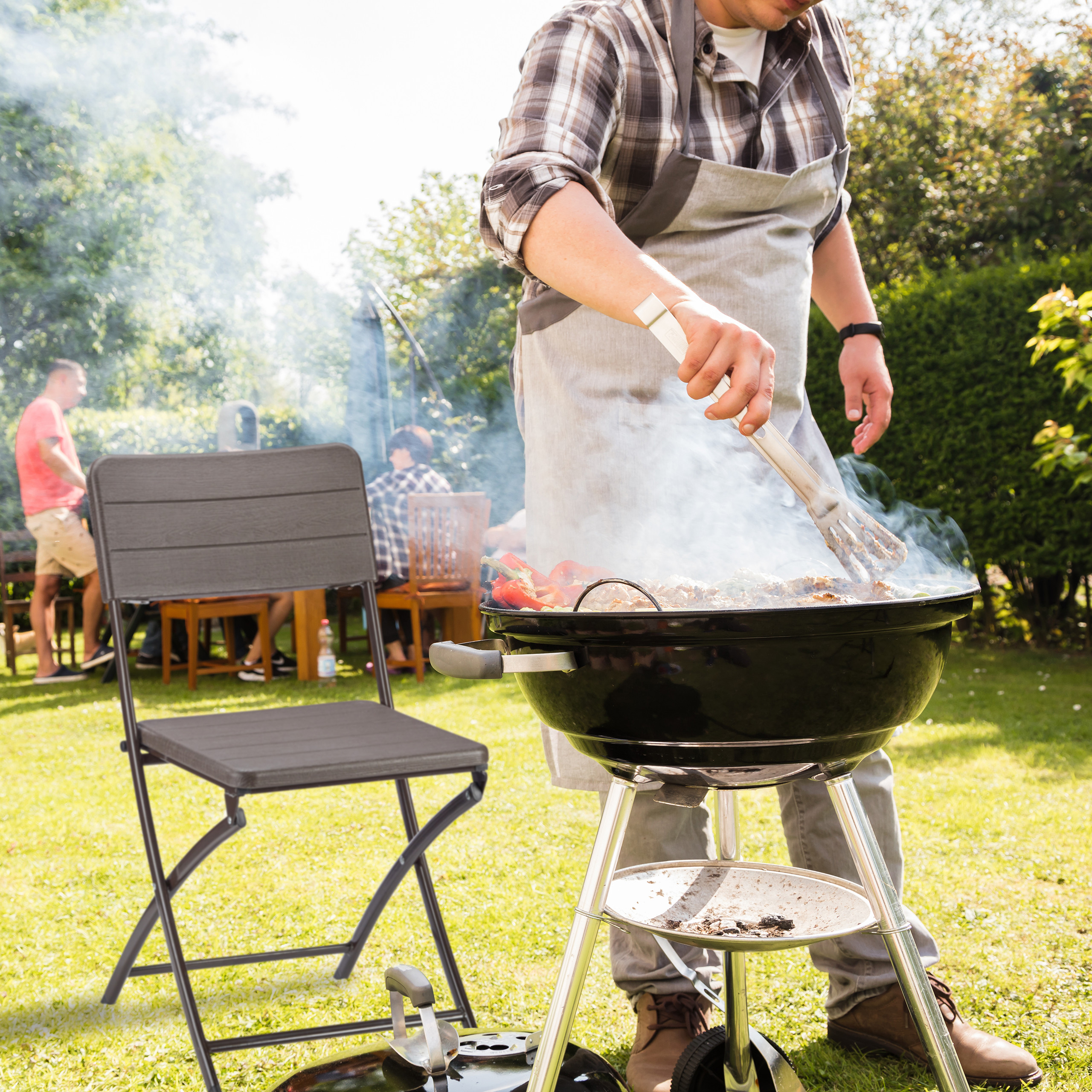 Ein Mann im grauen Apron grillt Fleisch im Garten mit einer grauen Stuhl im Vordergrund.