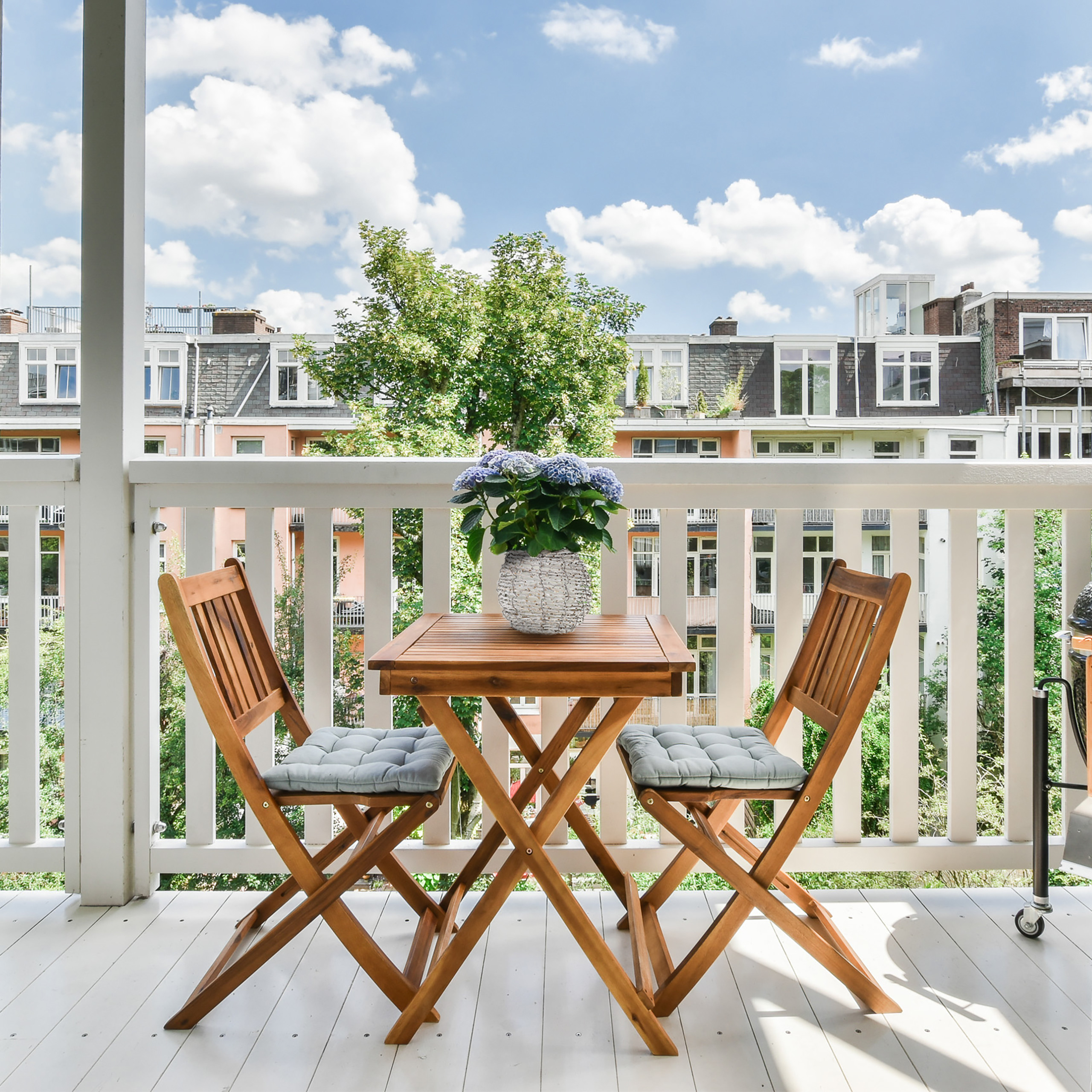 Ein Holztisch mit blauen Blumen und zwei Stühlen auf einem sonnigen Balkon mit Blick auf die Stadt.