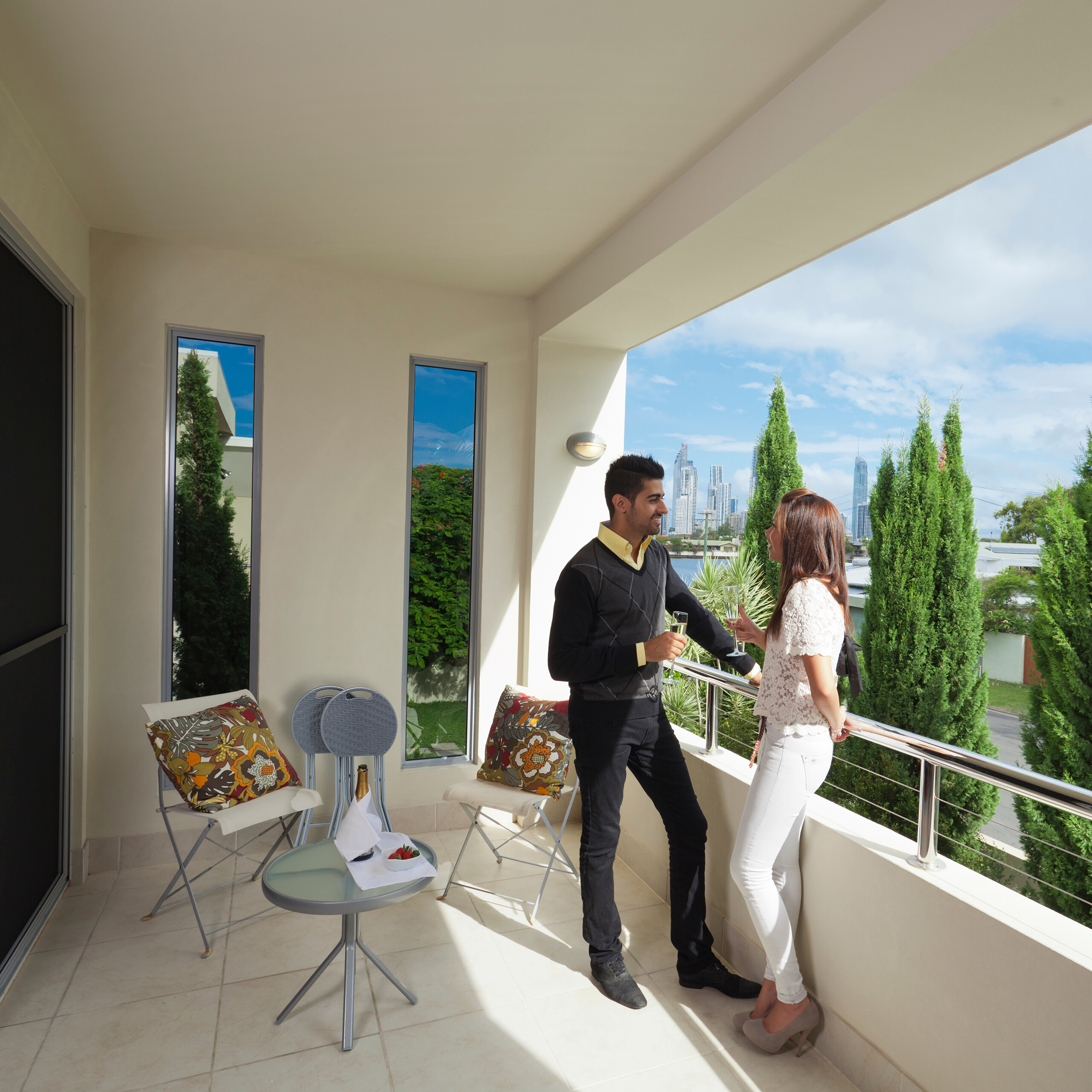 Ein lächelndes Paar auf einem sonnigen Balkon mit Getränken und Blick auf die Skyline der Stadt.