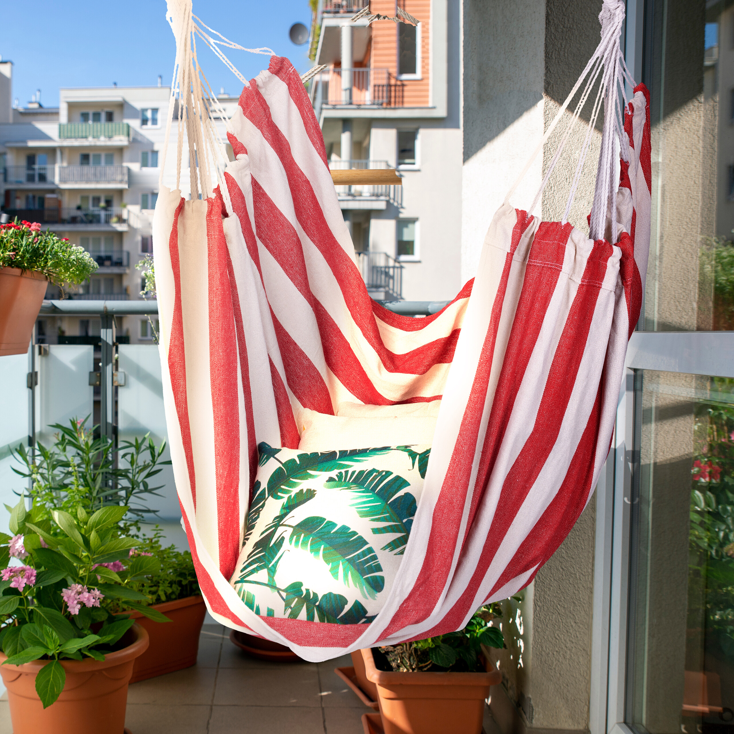 Rot-weiß gestreifter Hängesessel mit Kissen auf einem sonnigen Balkon mit Pflanzen.