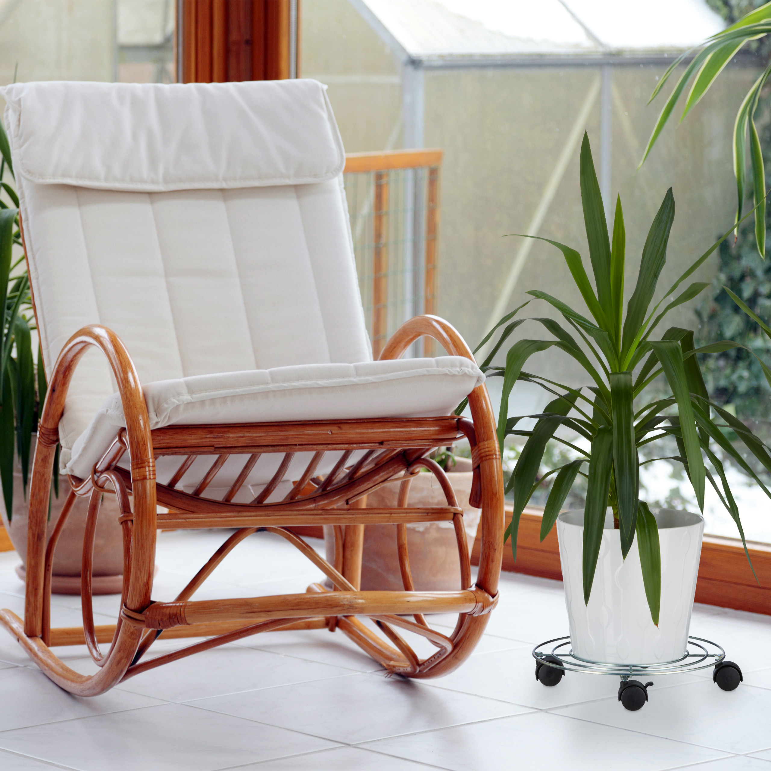 Ein weißer Schaukelstuhl aus Rattan steht auf Fliesen neben einer Grünpflanze in einem Topf mit Rollen.