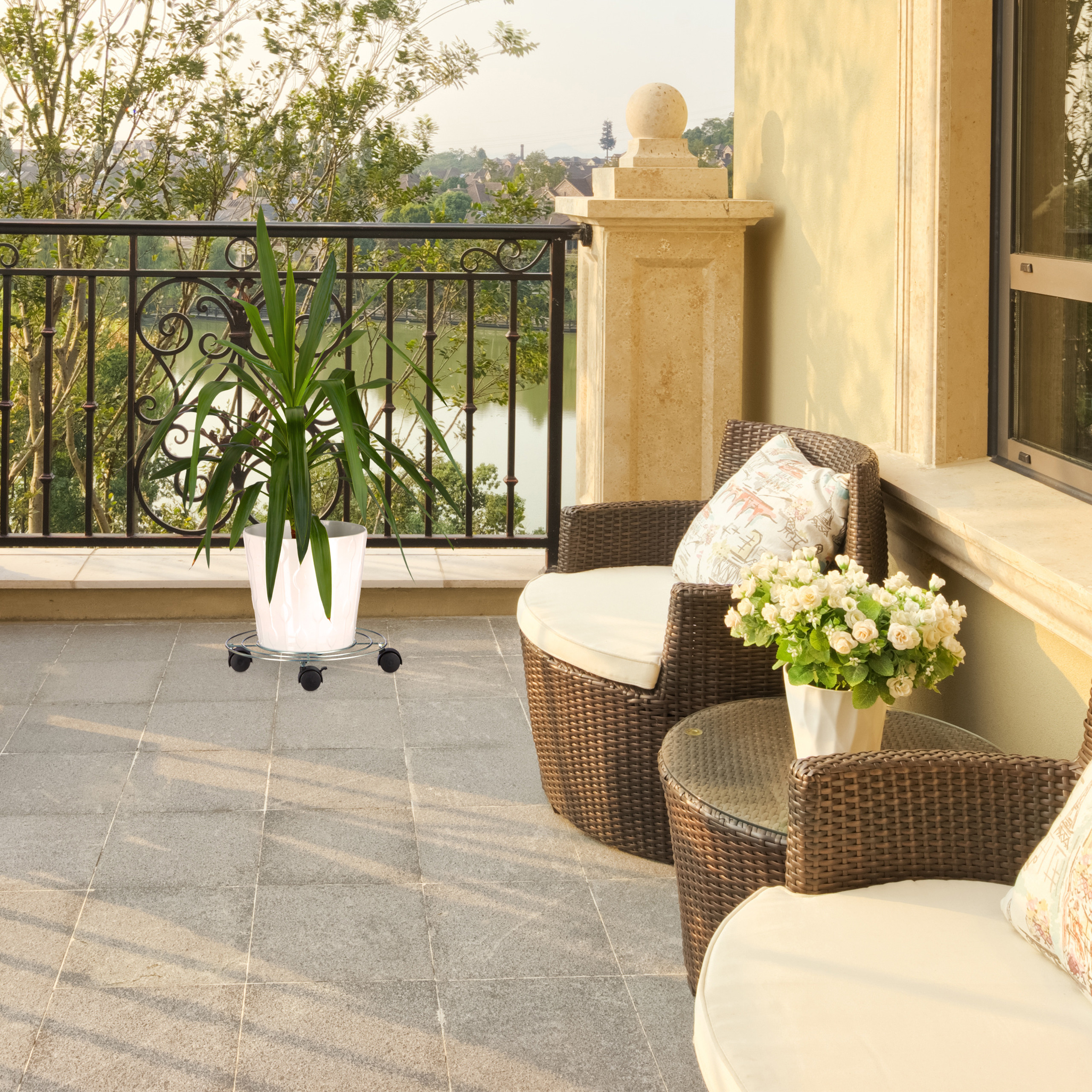 Ein sonniger Balkon mit Rattansesseln, einem Tisch mit Blumen, einer Topfpflanze auf Rädern und Blick auf einen See.