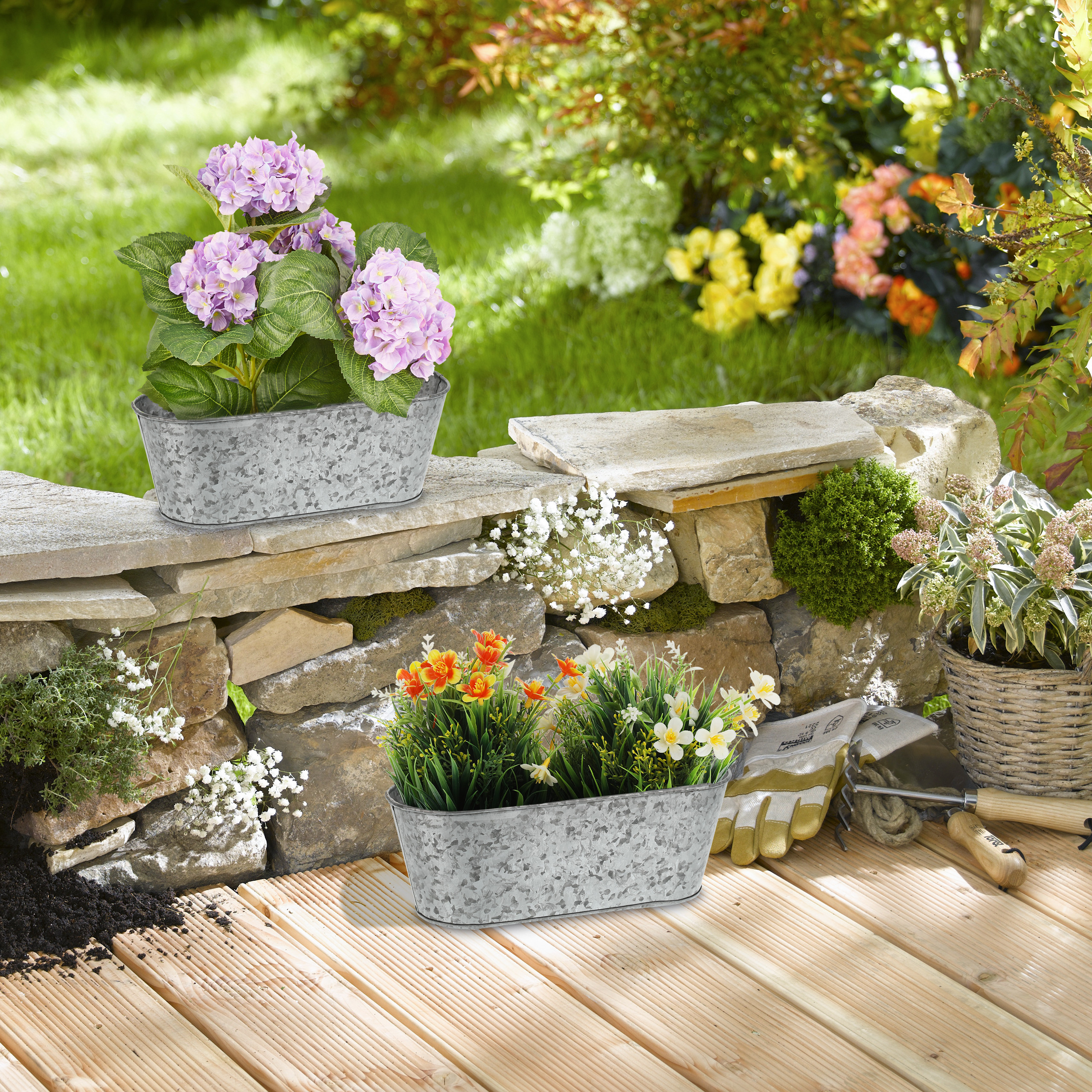 Zwei graue Metalltöpfe mit Blumen stehen auf einer Holzterrasse vor einer Steinmauer und einem grünen Garten.