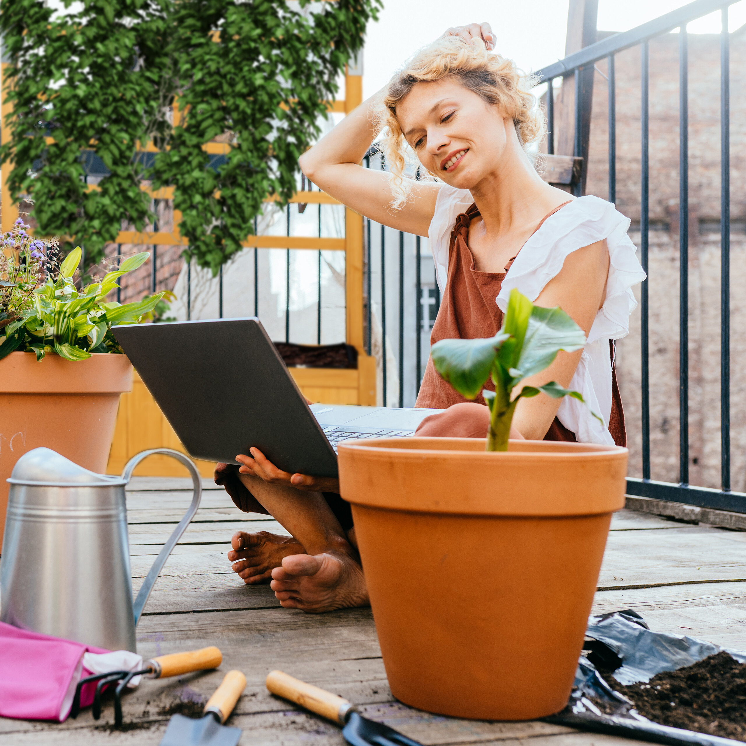 Eine blonde Frau sitzt auf einer Terrasse mit einem Laptop, umgeben von Pflanzen und Gartenwerkzeug.
