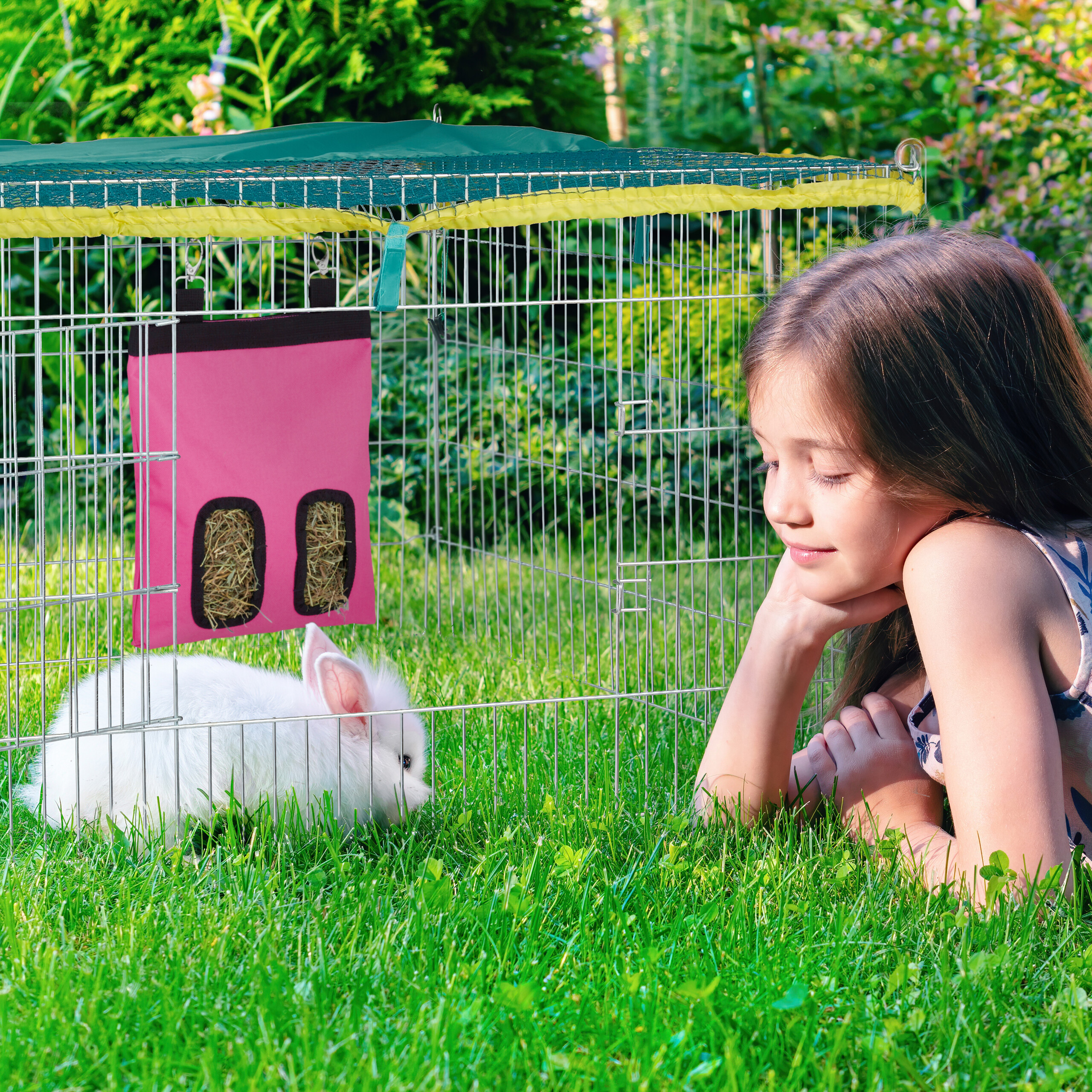 Ein junges Mädchen liegt im Gras und beobachtet ein weißes Kaninchen in einem drahtenen Käfig mit grünem Dach.