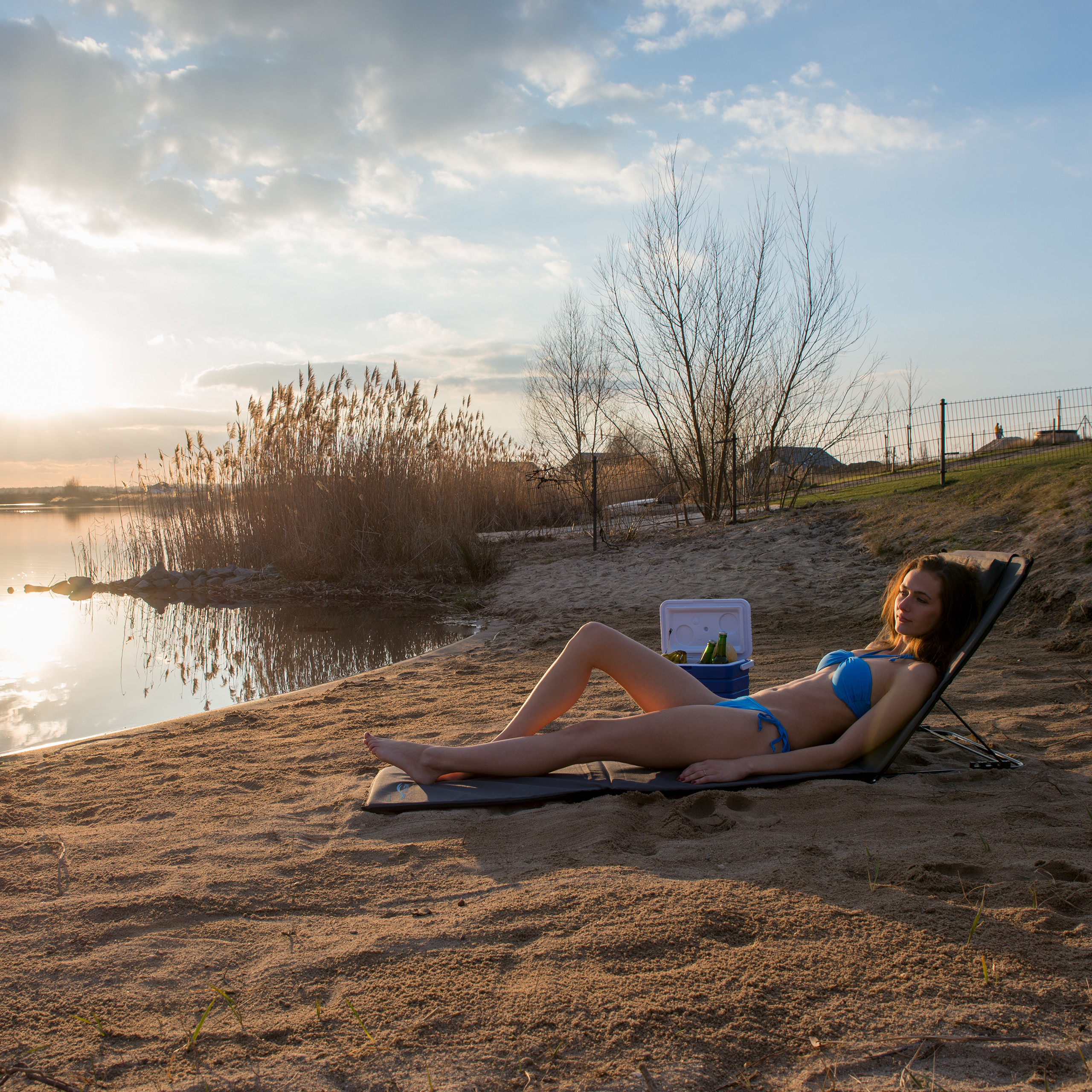 Eine Frau in blauem Bikini entspannt am sandigen Seeufer bei Sonnenuntergang neben einer Kühlbox.