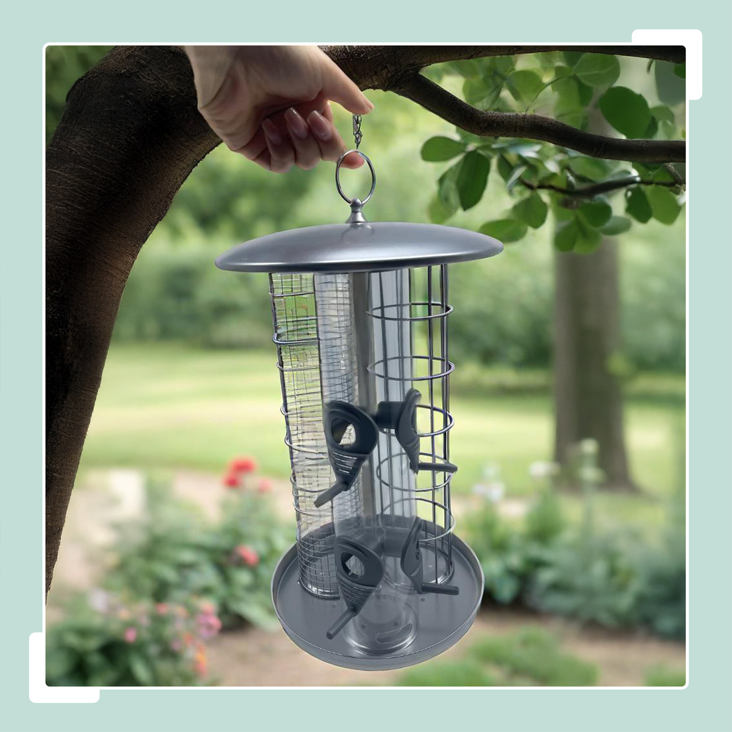 Eine Hand hält einen grauen Vogelfutterspender aus Metall an einem Ast in einem Garten.