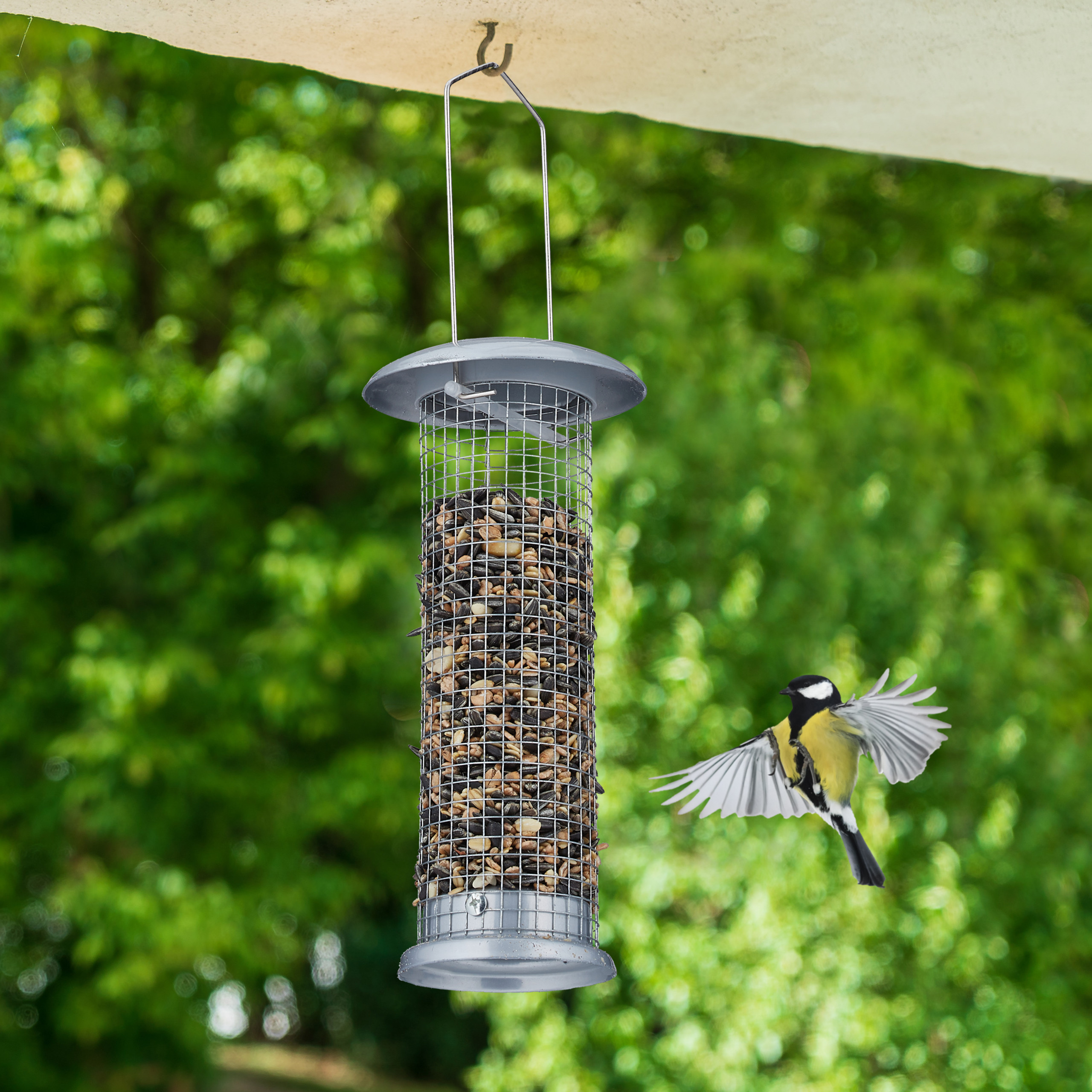 Eine Kohlmeise fliegt auf ein hängendes Vogelfutterhaus mit Samen zu, vor grünem Hintergrund.