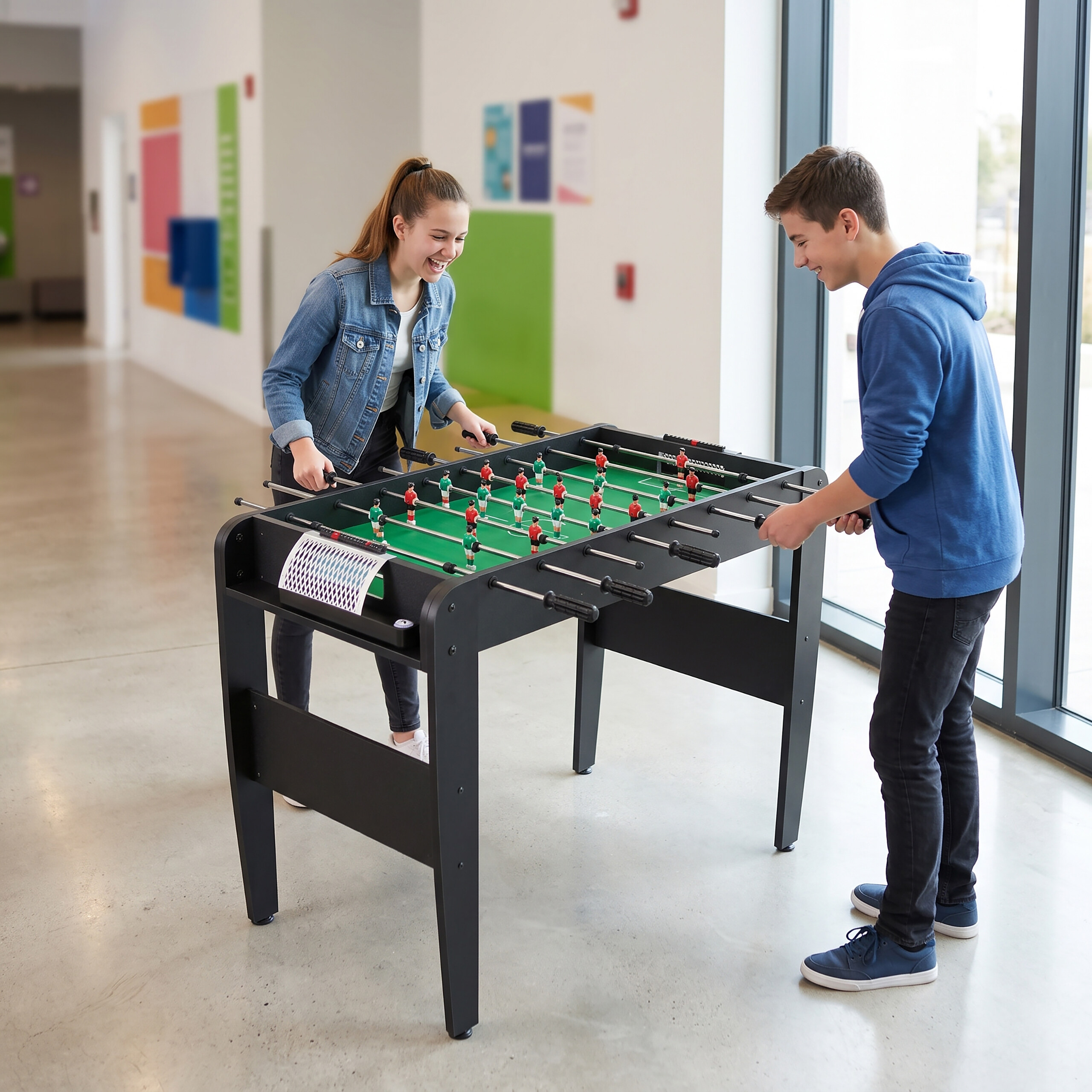 Ein lächelnder Junge und ein Mädchen spielen Tischfußball in einem hellen Flur an großen Fenstern.