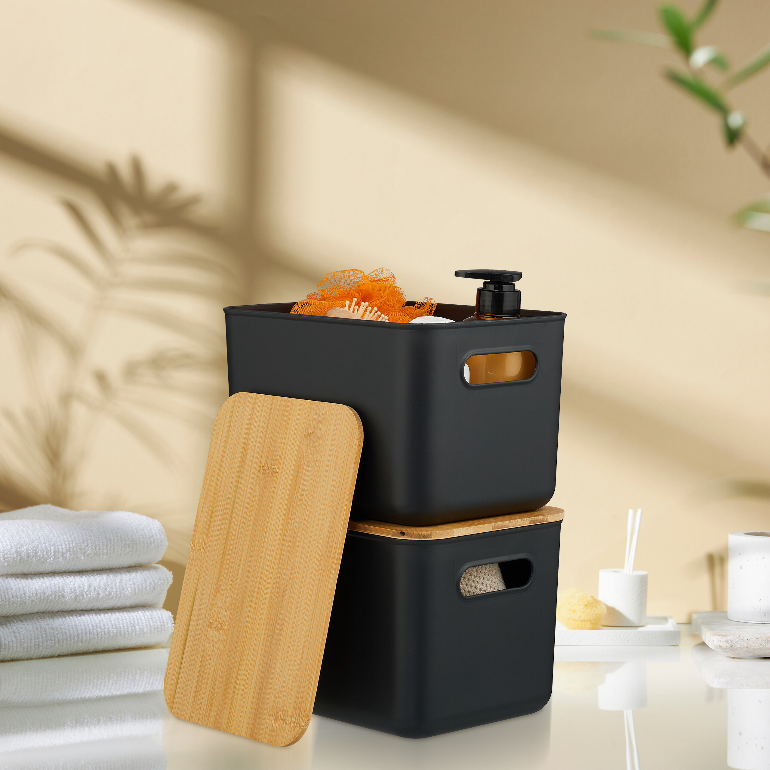 Zwei schwarze Aufbewahrungskörbe mit Holzdeckeln auf einer Oberfläche, neben weißen Handtüchern und Badaccessoires.
