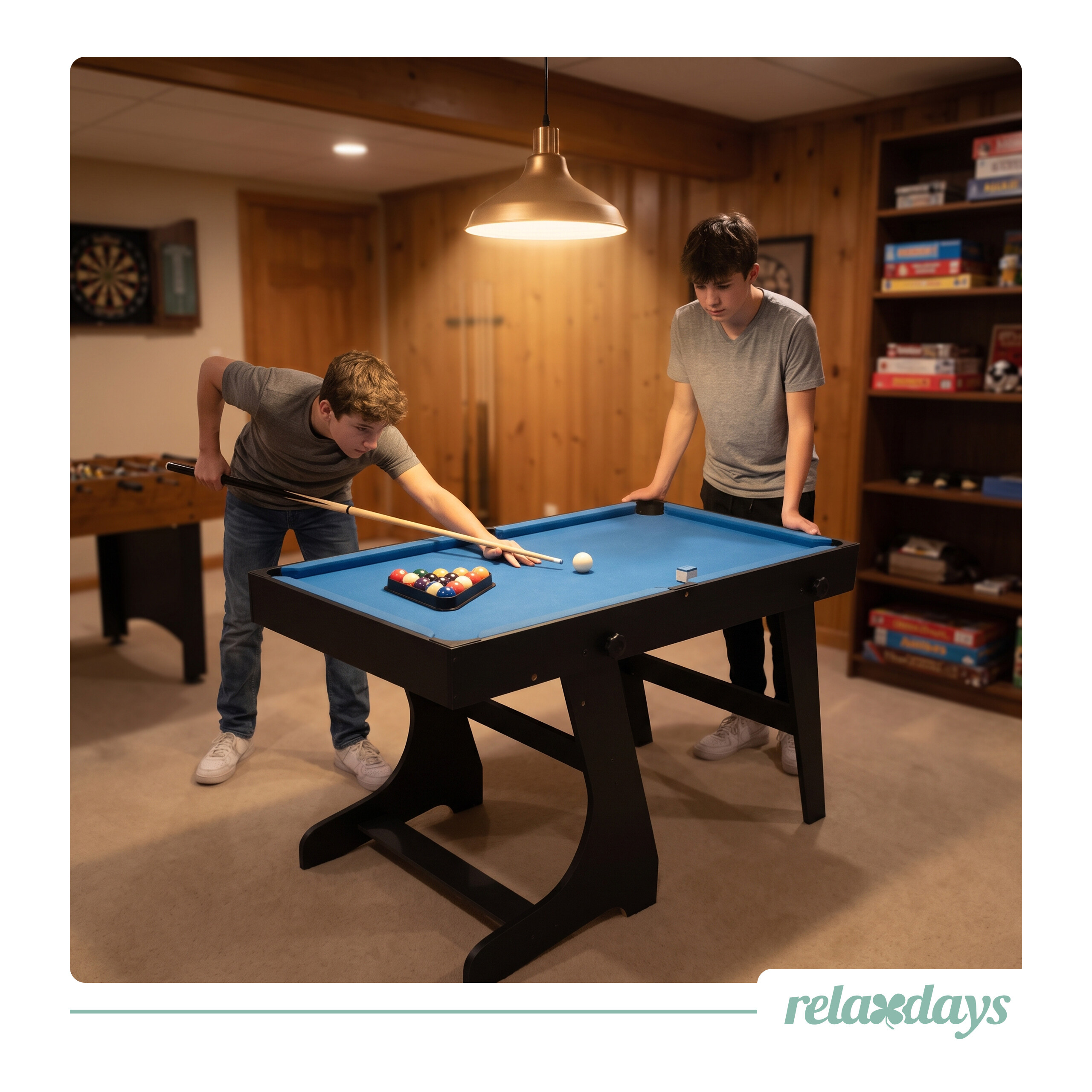 Zwei Jungen spielen Billard in einem Keller mit Holzwänden, einem blauen Billardtisch und Hängelampe.