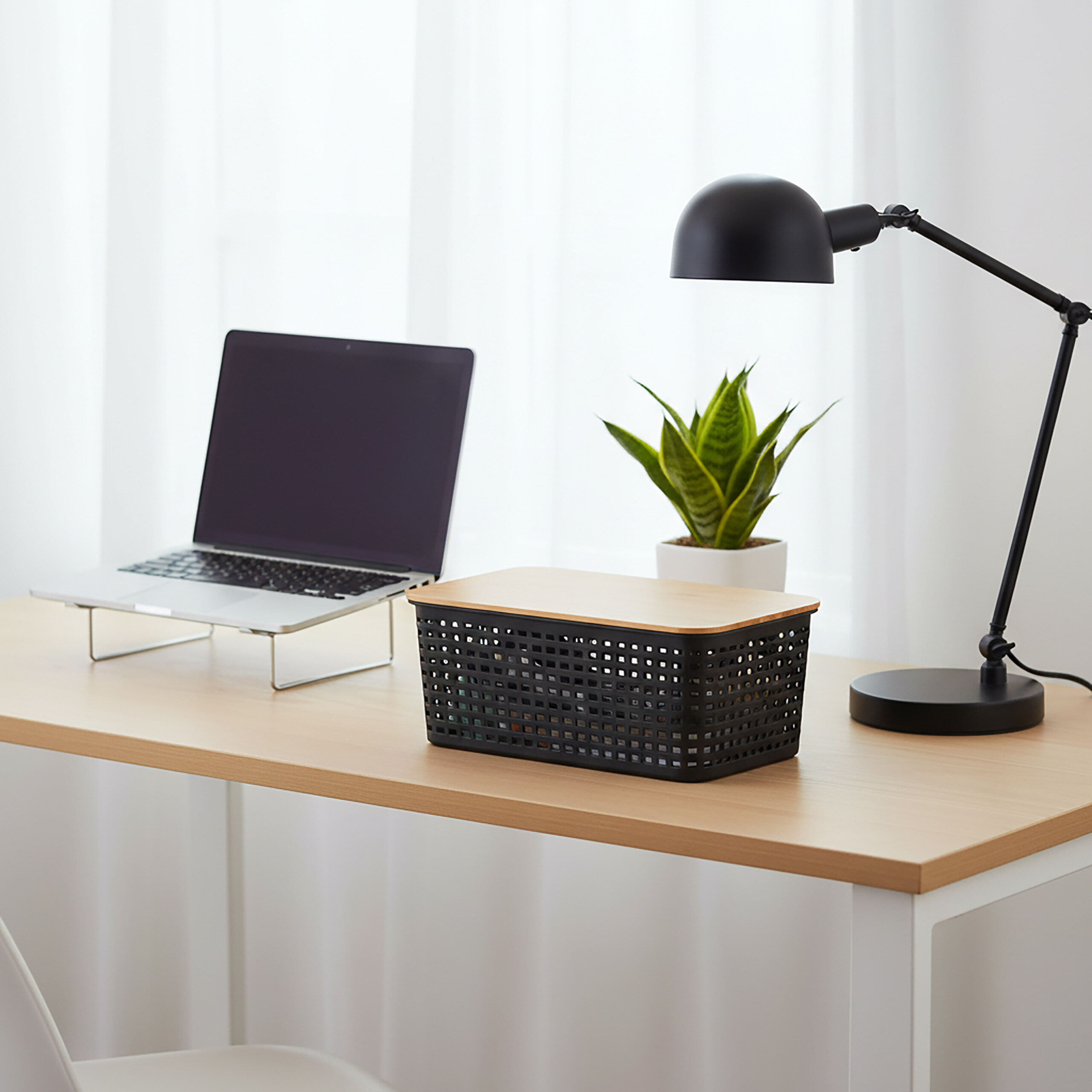 Ein Holztisch zeigt einen Laptop auf einem Ständer, einen schwarzen Korb mit Holzdeckel, eine Topfpflanze und eine Schreibtischlampe.