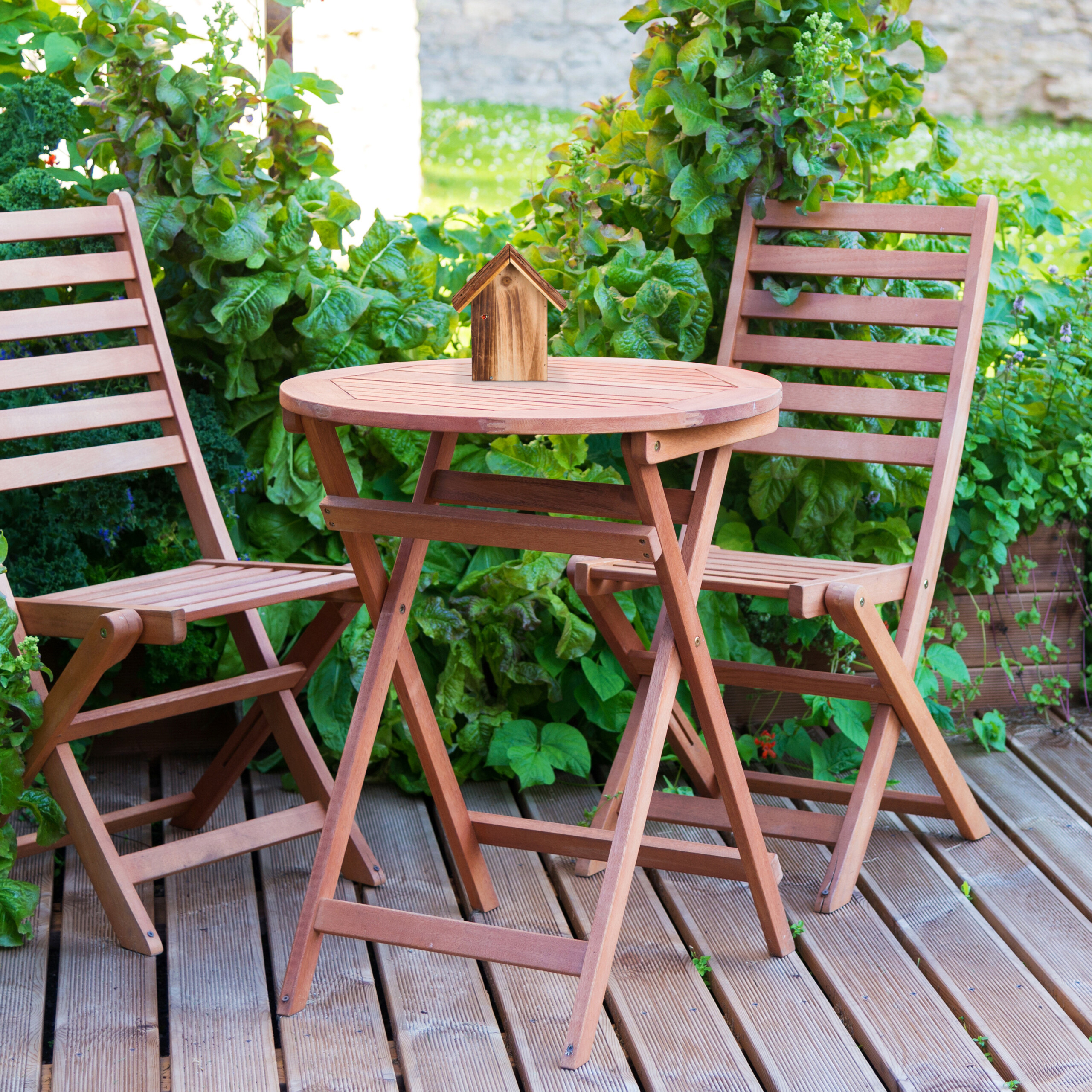 Ein kleiner Holztisch mit Vogelhäuschen und zwei Stühlen auf einer Holzterrasse im Garten.