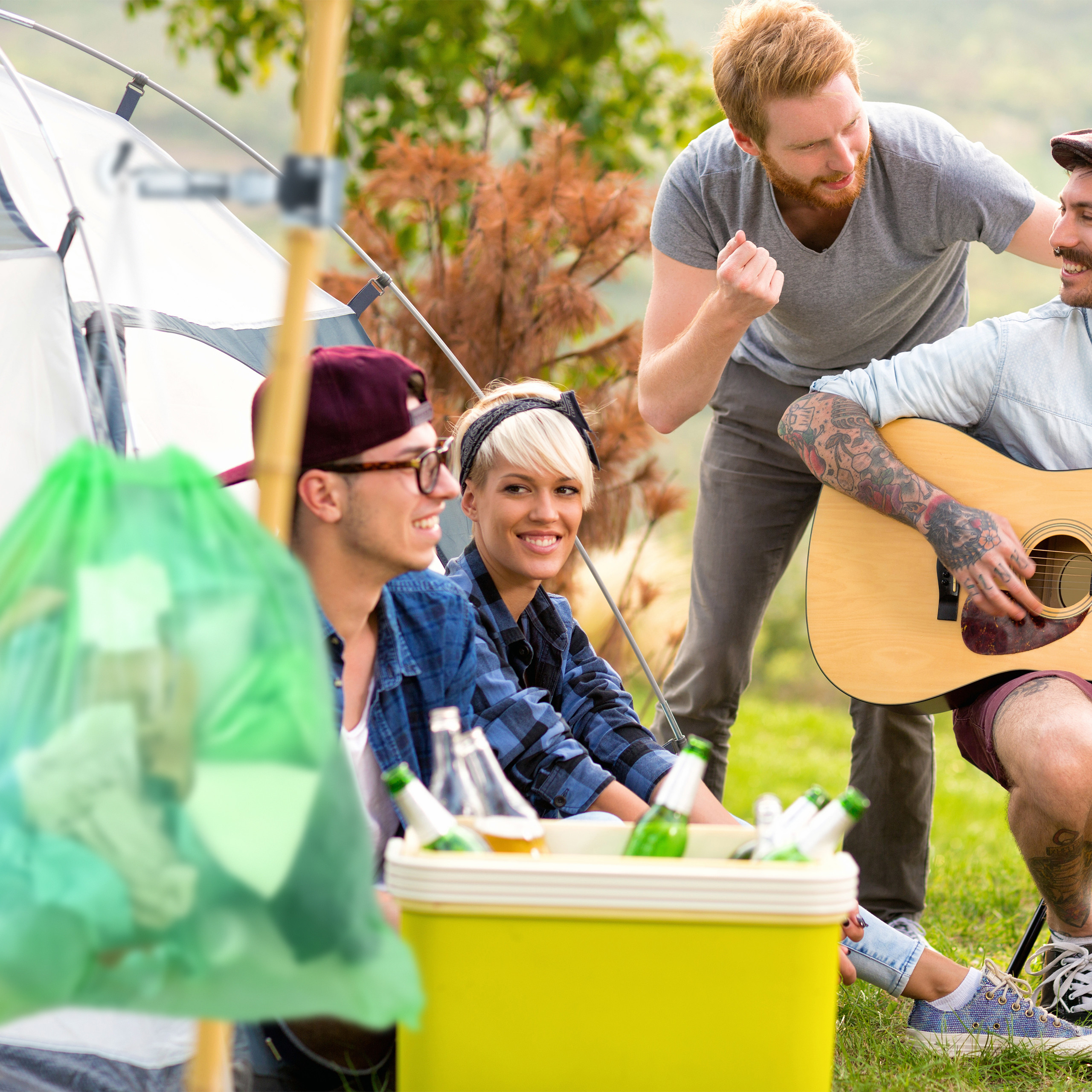Vier Freunde campen in der Natur, einer spielt Gitarre, daneben steht eine Kühlbox mit Getränken.