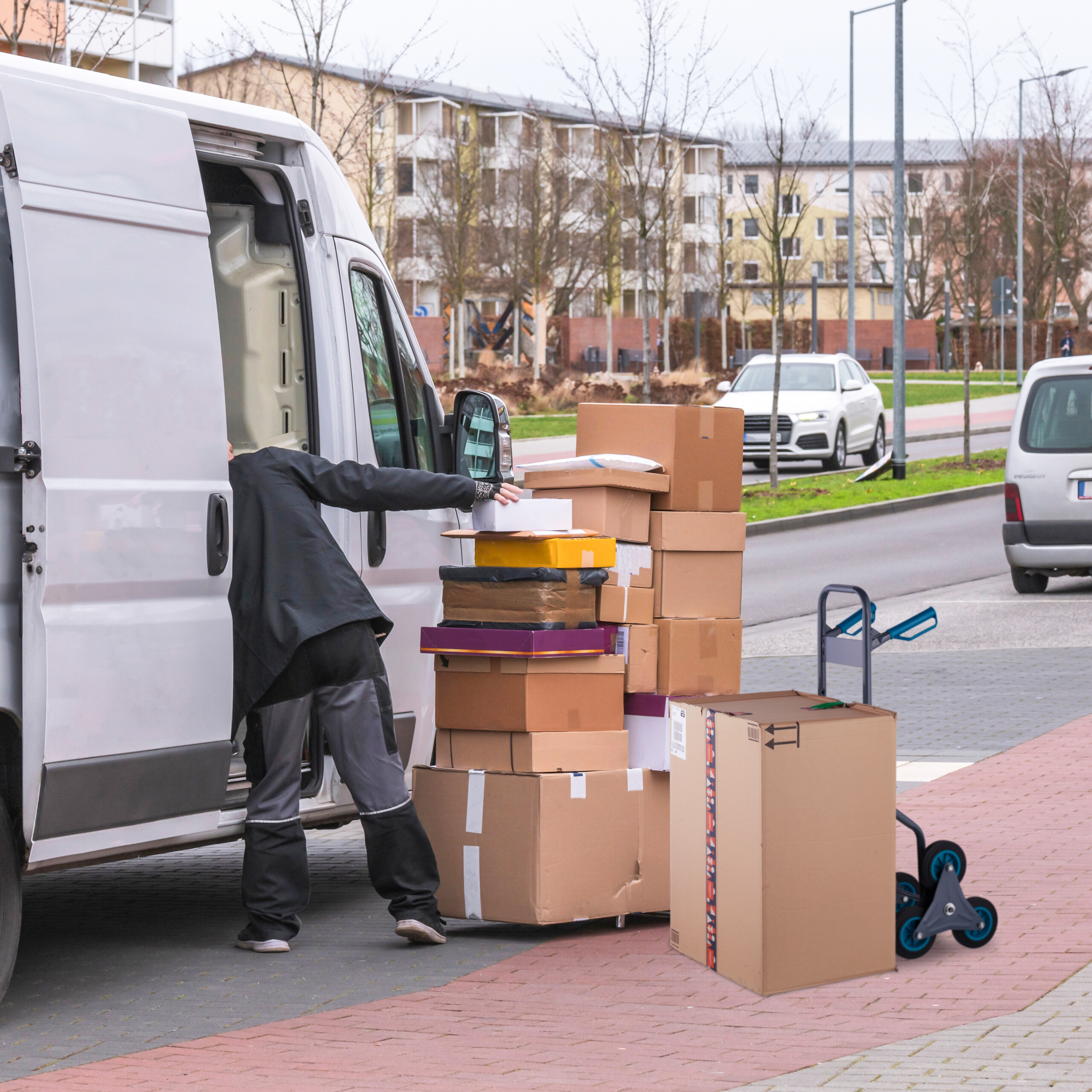 Ein Lieferant in schwarzer Kleidung lädt Pakete aus einem weißen Transporter auf einem städtischen Parkplatz aus.