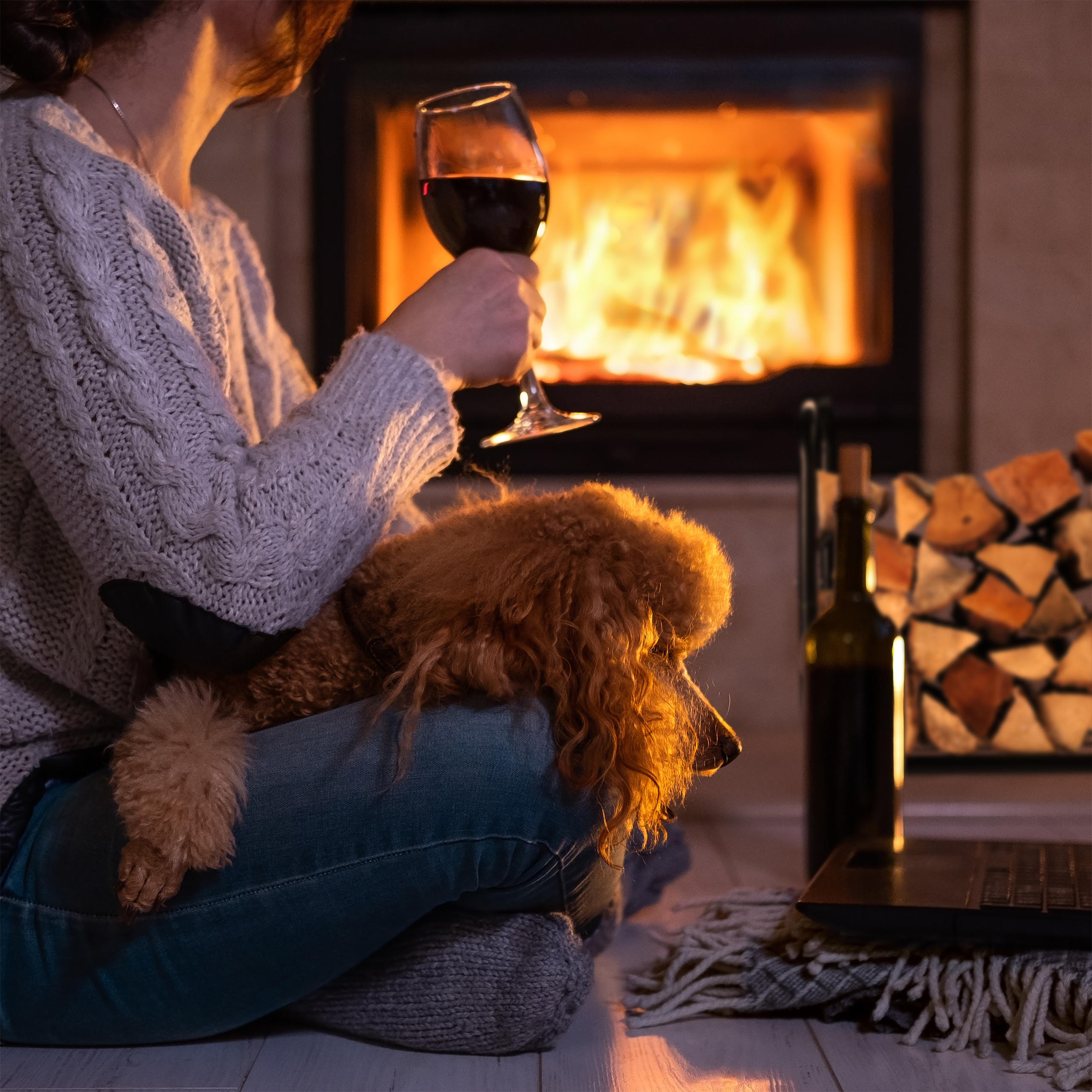 Frau mit Pulli hält Rotwein, ein Hund sitzt auf ihrem Schoß vor Kamin.