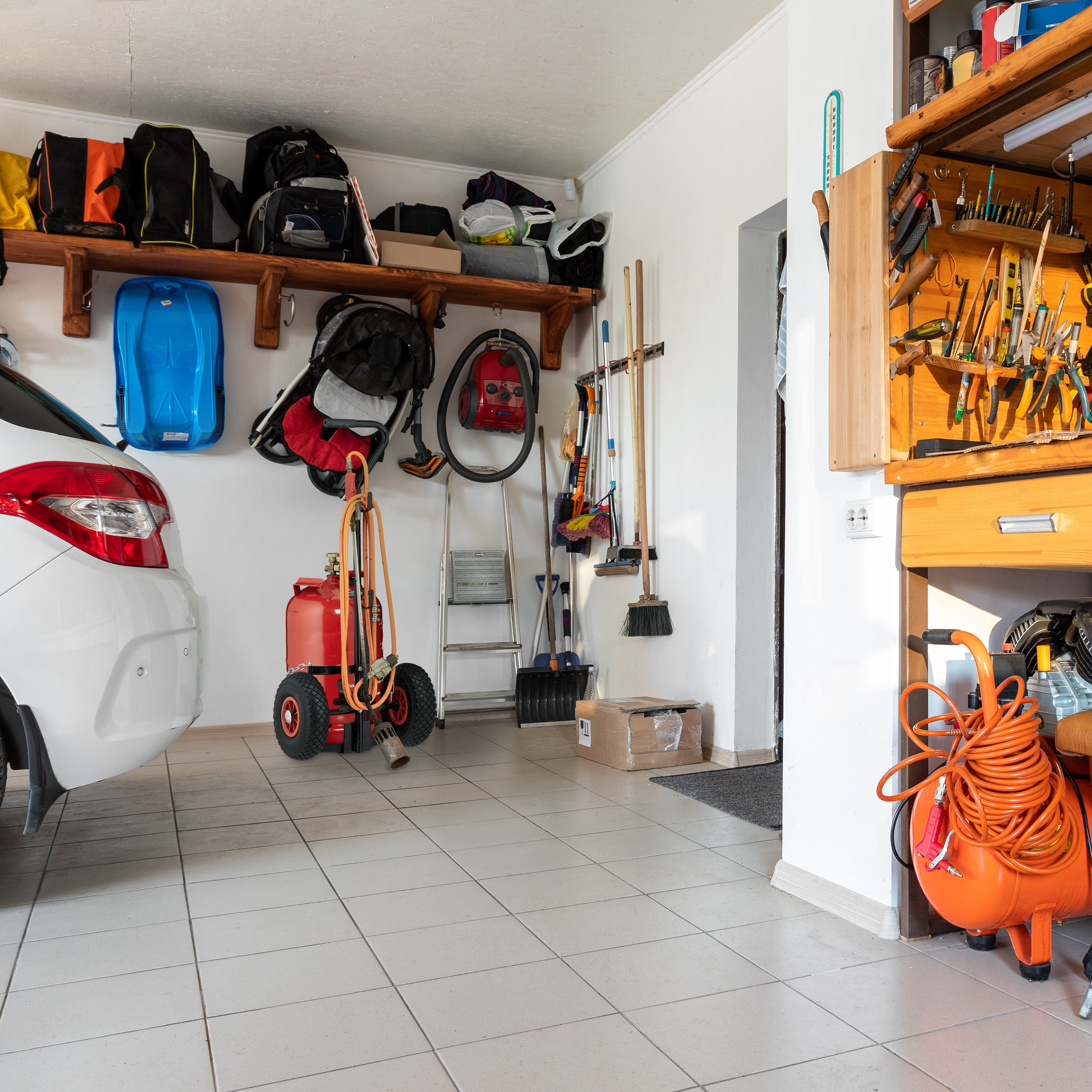 Ordentliche Garage mit weißem Auto, Werkzeugregal, rotem Kompressor, Leiter und aufgehängten Taschen.