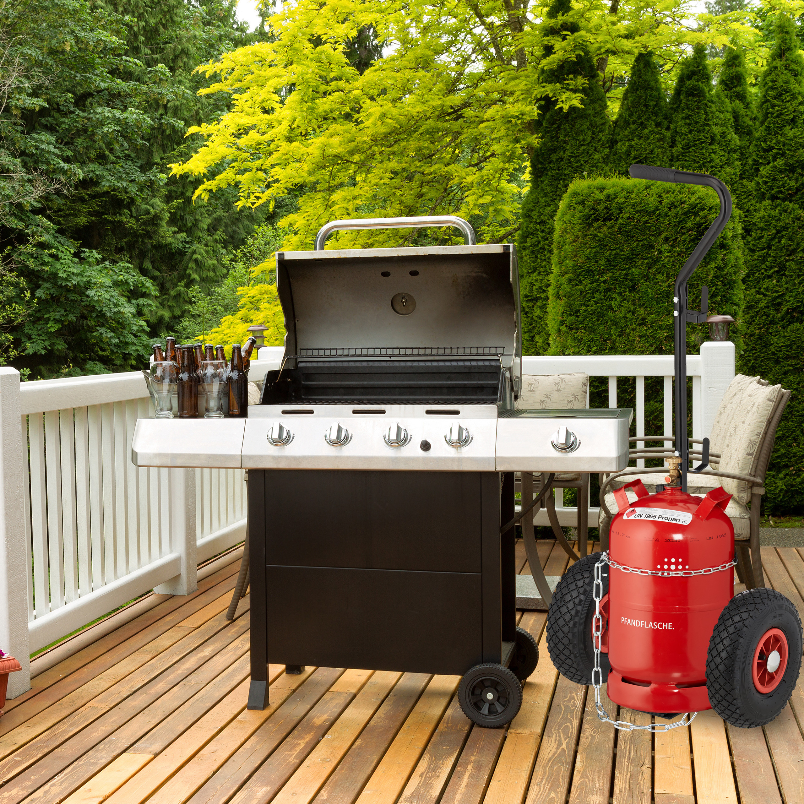 Ein offener Gasgrill auf einer Holzterrasse mit Flaschen und einer roten Propangasflasche auf Rädern daneben.