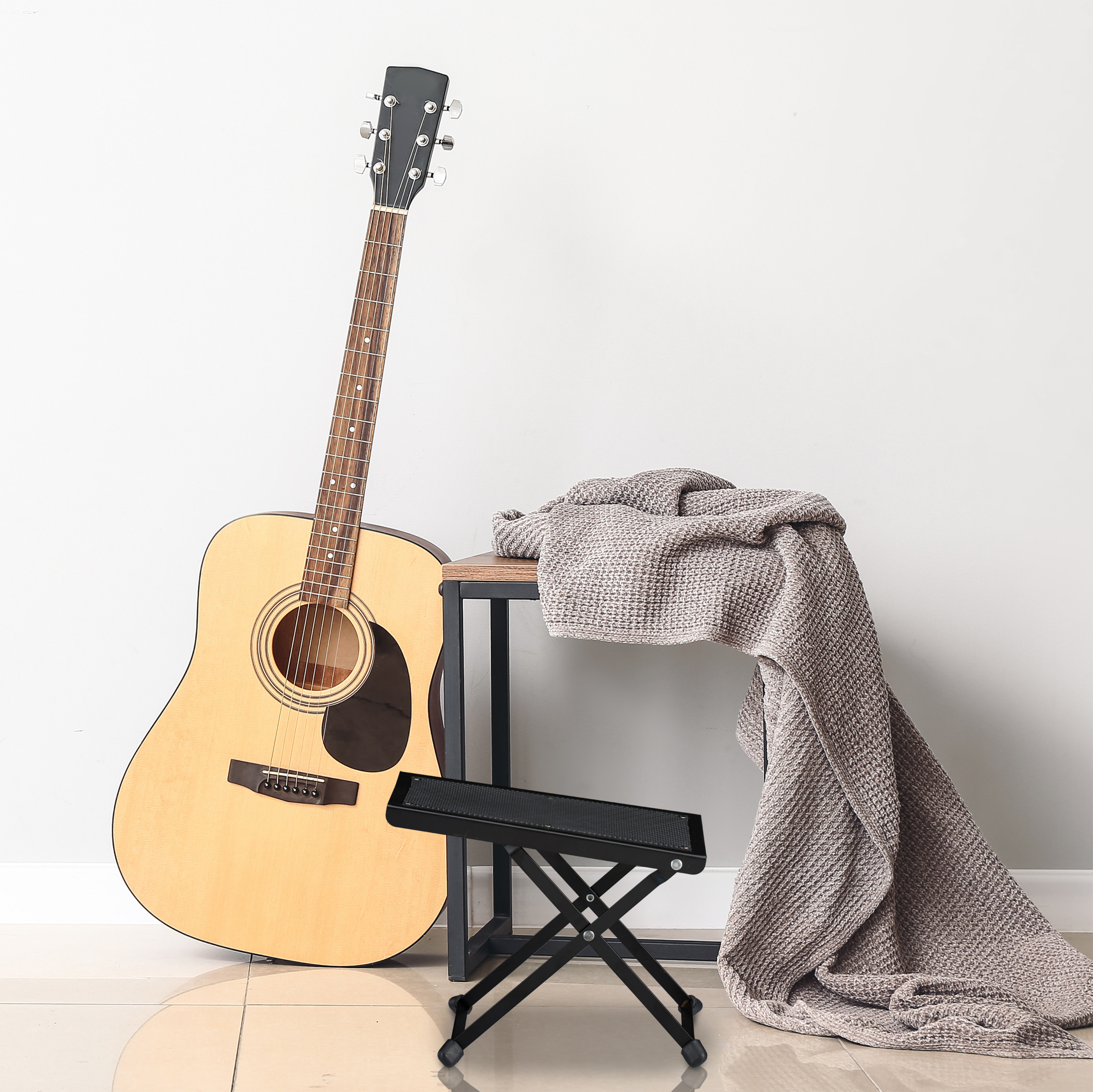 Eine Akustikgitarre lehnt an einer Wand neben einem kleinen Tisch mit einer grauen Decke und einem Hocker.