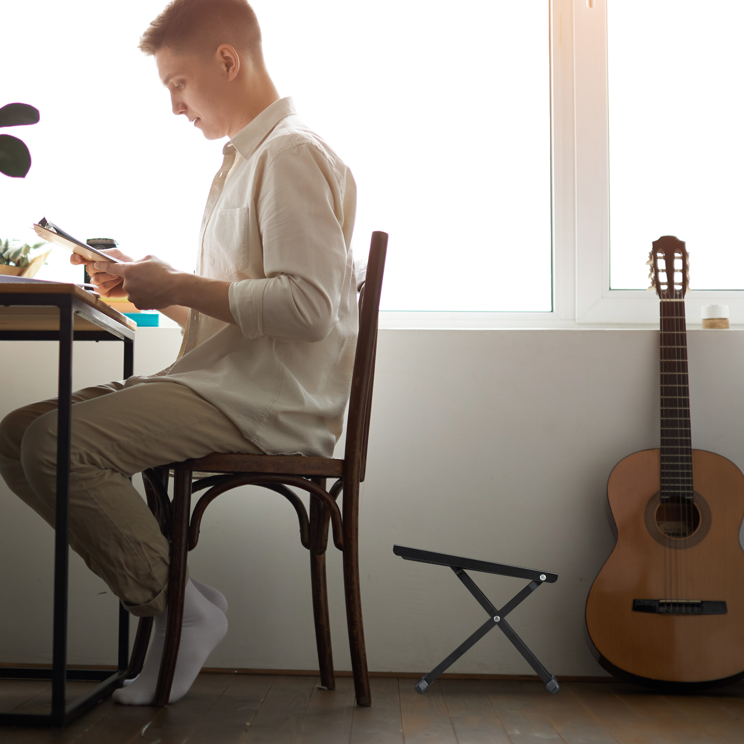 Ein junger Mann sitzt am Holztisch vor einem Fenster und liest Dokumente. Eine Akustikgitarre steht an der Wand.