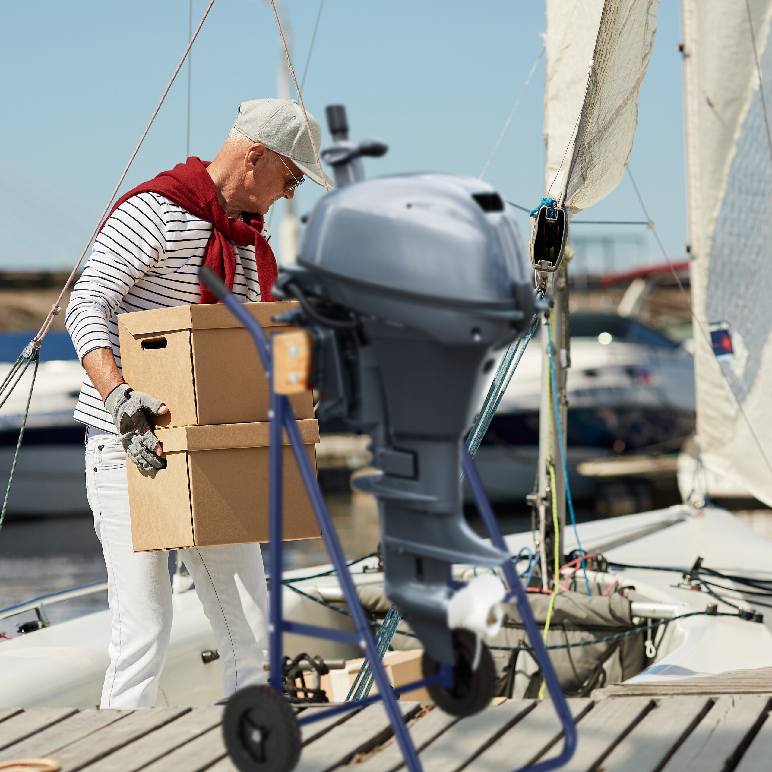 Älterer Mann in gestreiftem Hemd trägt Pappkartons auf Segelbootsteg mit Bootsmotor im Vordergrund.