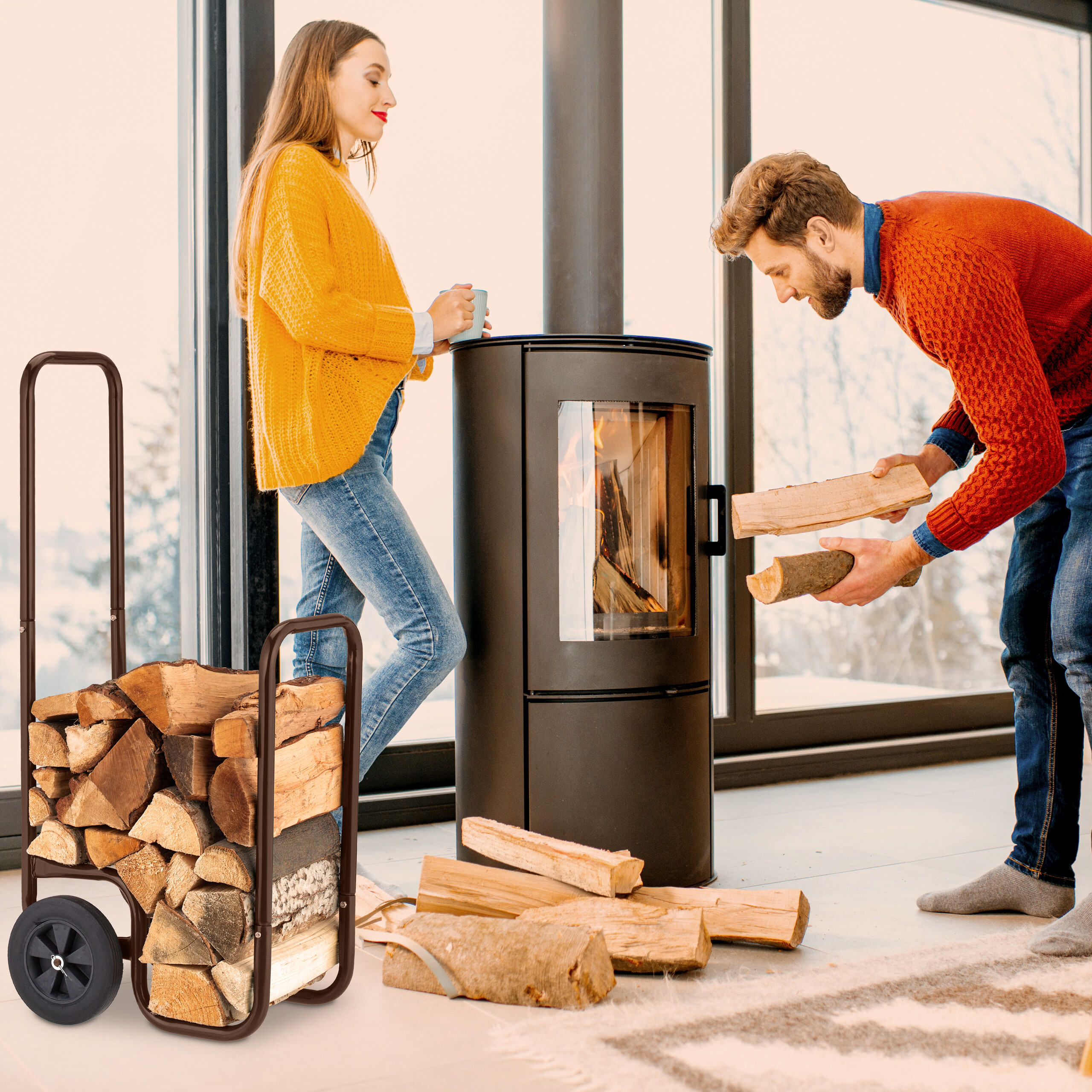 Ein Mann legt Holz in einen Ofen, während eine Frau mit Tasse zusieht, im Vordergrund ein Holzkarren.
