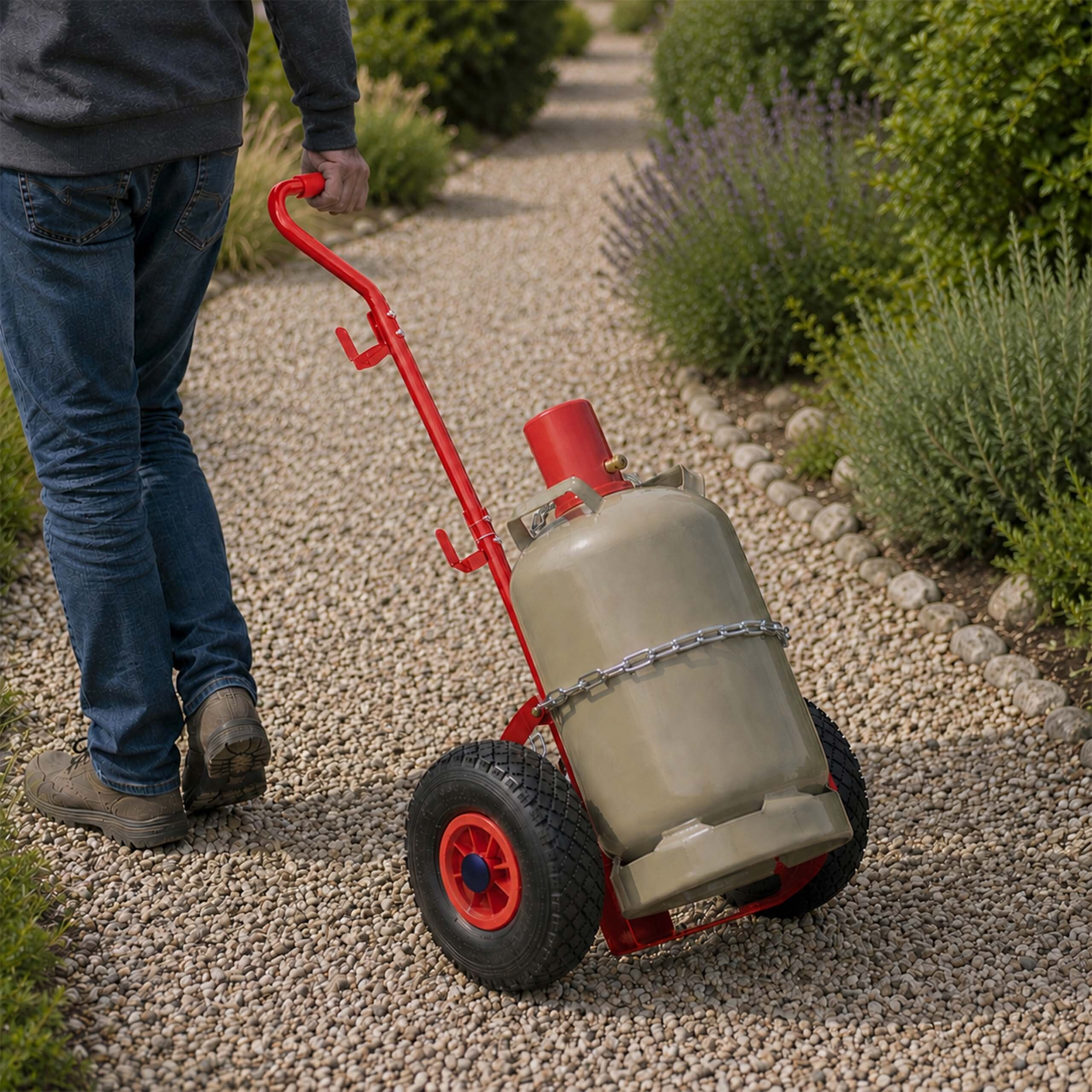 Ein Mann zieht einen roten Transportwagen mit einer großen Gasflasche auf einem Kiesweg im Garten.