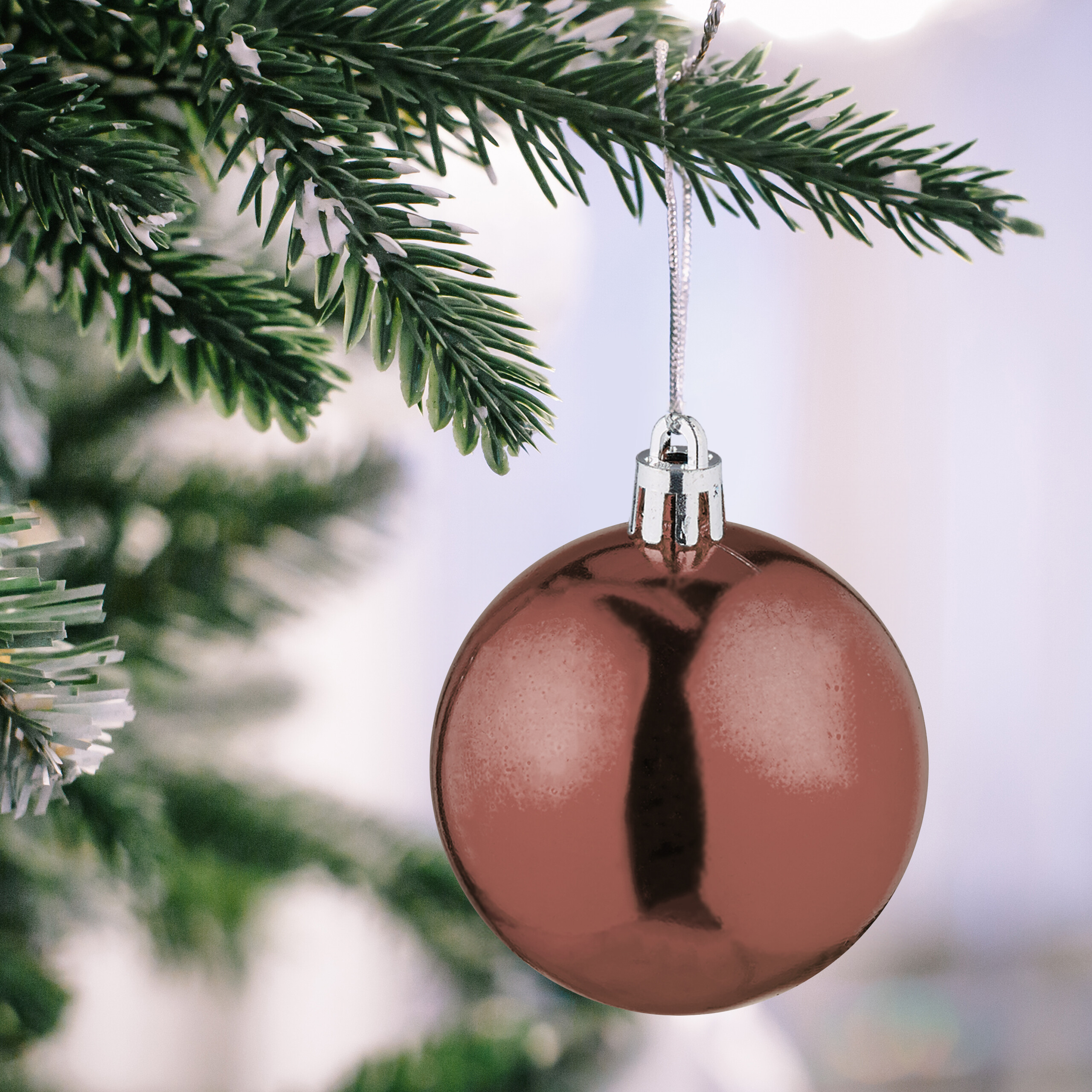 Eine glänzende kupferfarbene Weihnachtskugel hängt an einem verschneiten Weihnachtsbaum.