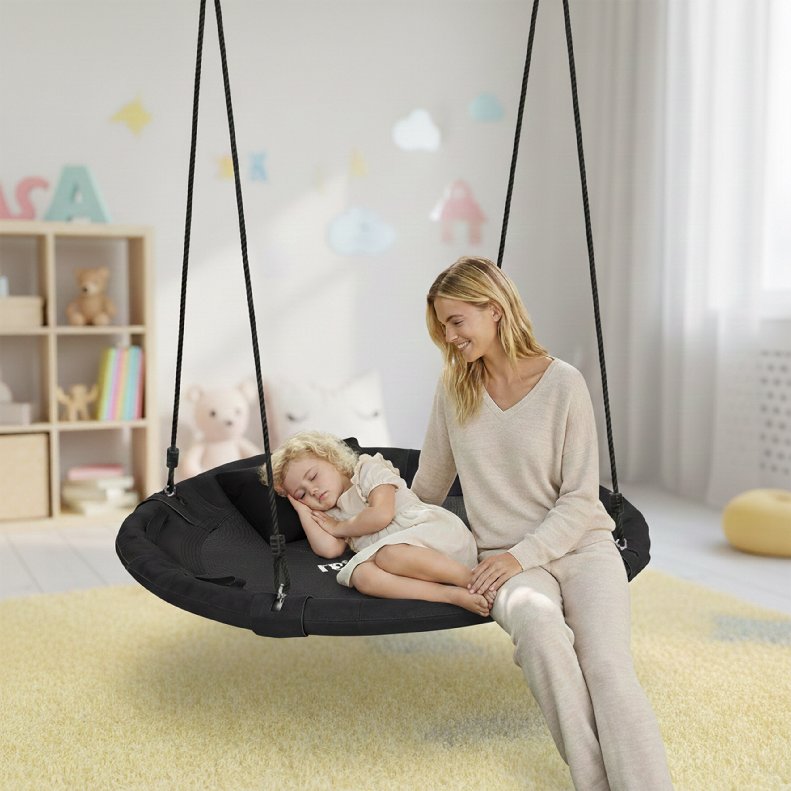 Eine lächelnde Frau sitzt auf einer schwarzen Schaukel, während ein Kind friedlich neben ihr in einem Spielzimmer schläft.