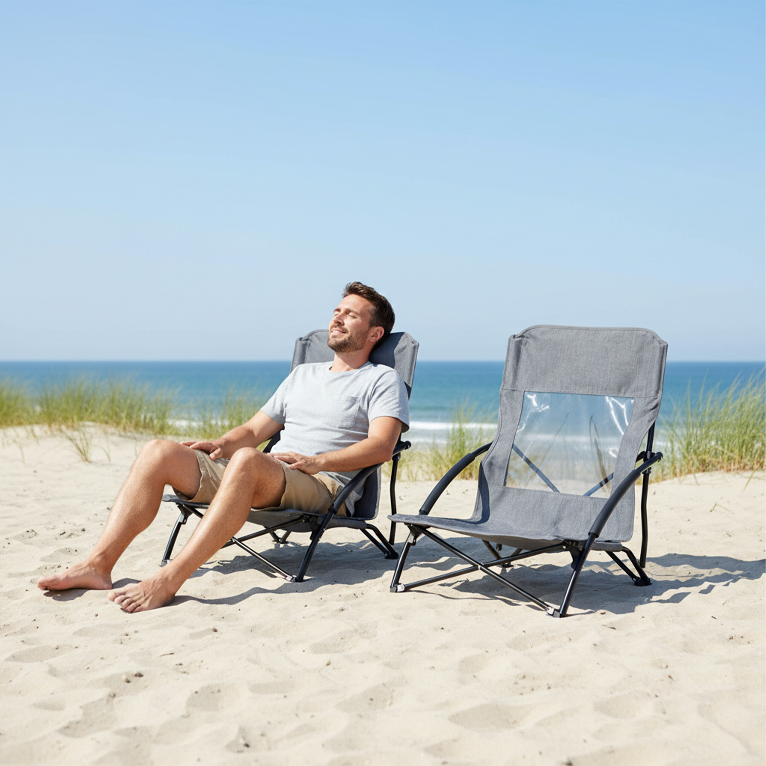 Ein Mann sitzt in einem grauen Strandstuhl und genießt die Sonne am sandigen Strand mit Blick auf das Meer.