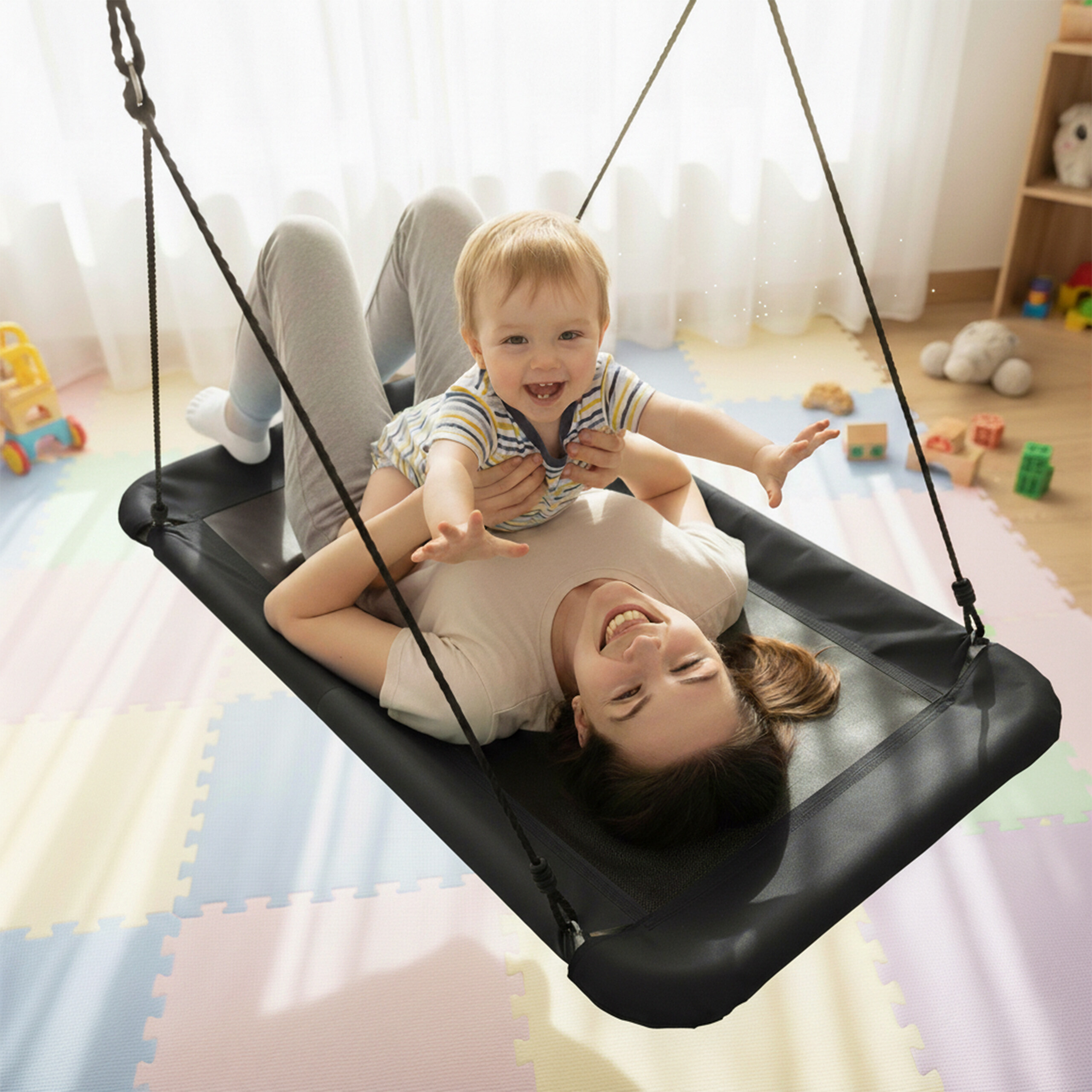 Eine glückliche Mutter und ihr Kleinkind lächeln, während sie zusammen auf einer schwarzen Hängematte im Kinderzimmer schaukeln.