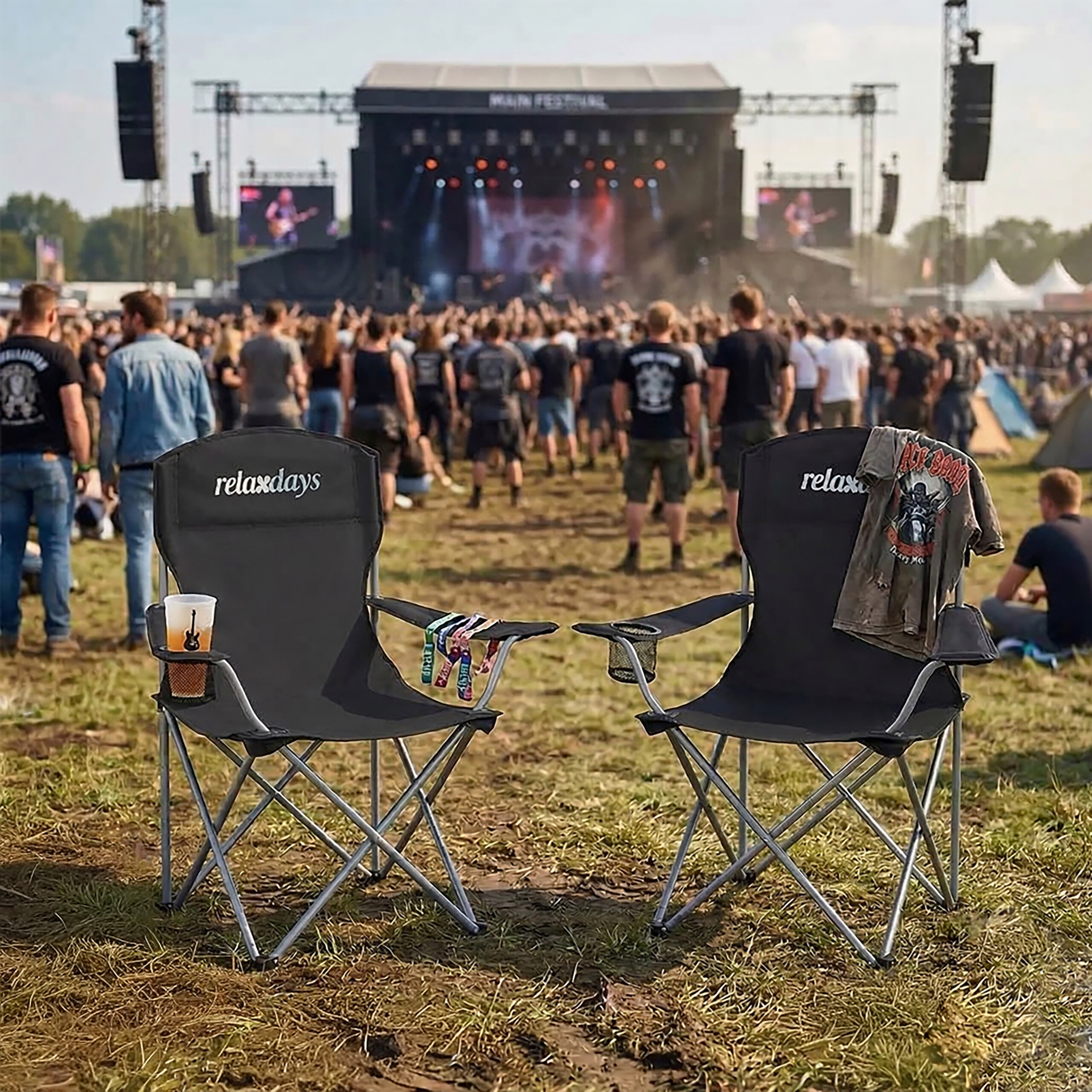 Zwei schwarze Campingstühle stehen auf einer Wiese vor einer Festivalbühne mit Publikum.