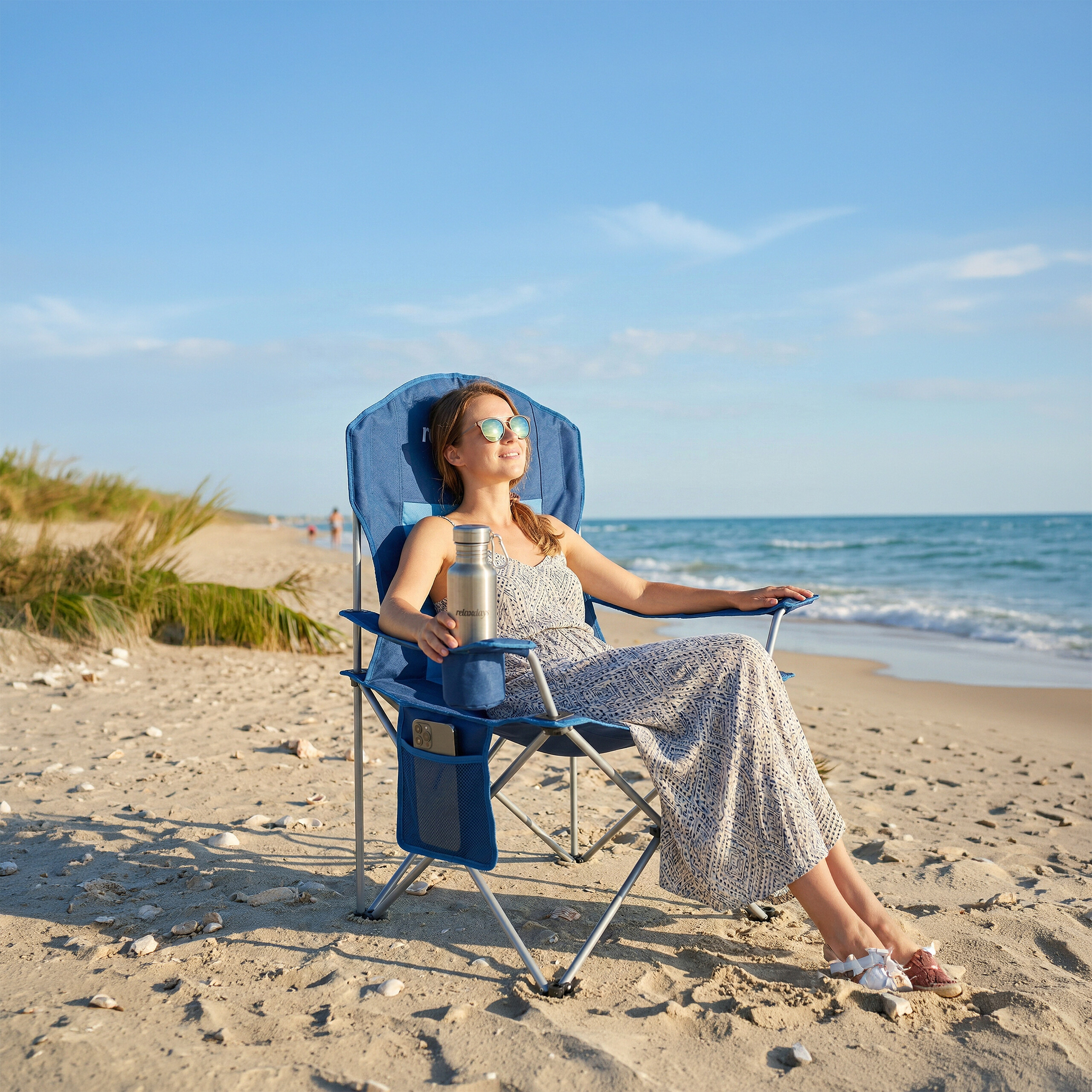 Eine Frau entspannt in einem blauen Klappstuhl an einem sonnigen Strand und hält eine silberne Wasserflasche.