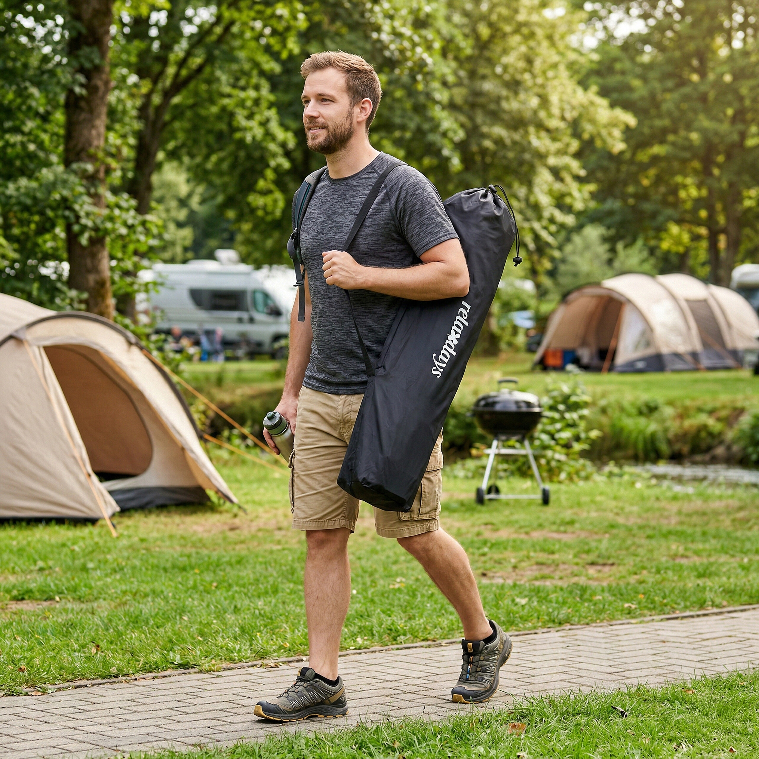 Ein junger Mann mit Bart geht auf einem Campingplatz auf einem Steinweg, trägt einen schwarzen Sack und ein Zelt im Hintergrund.